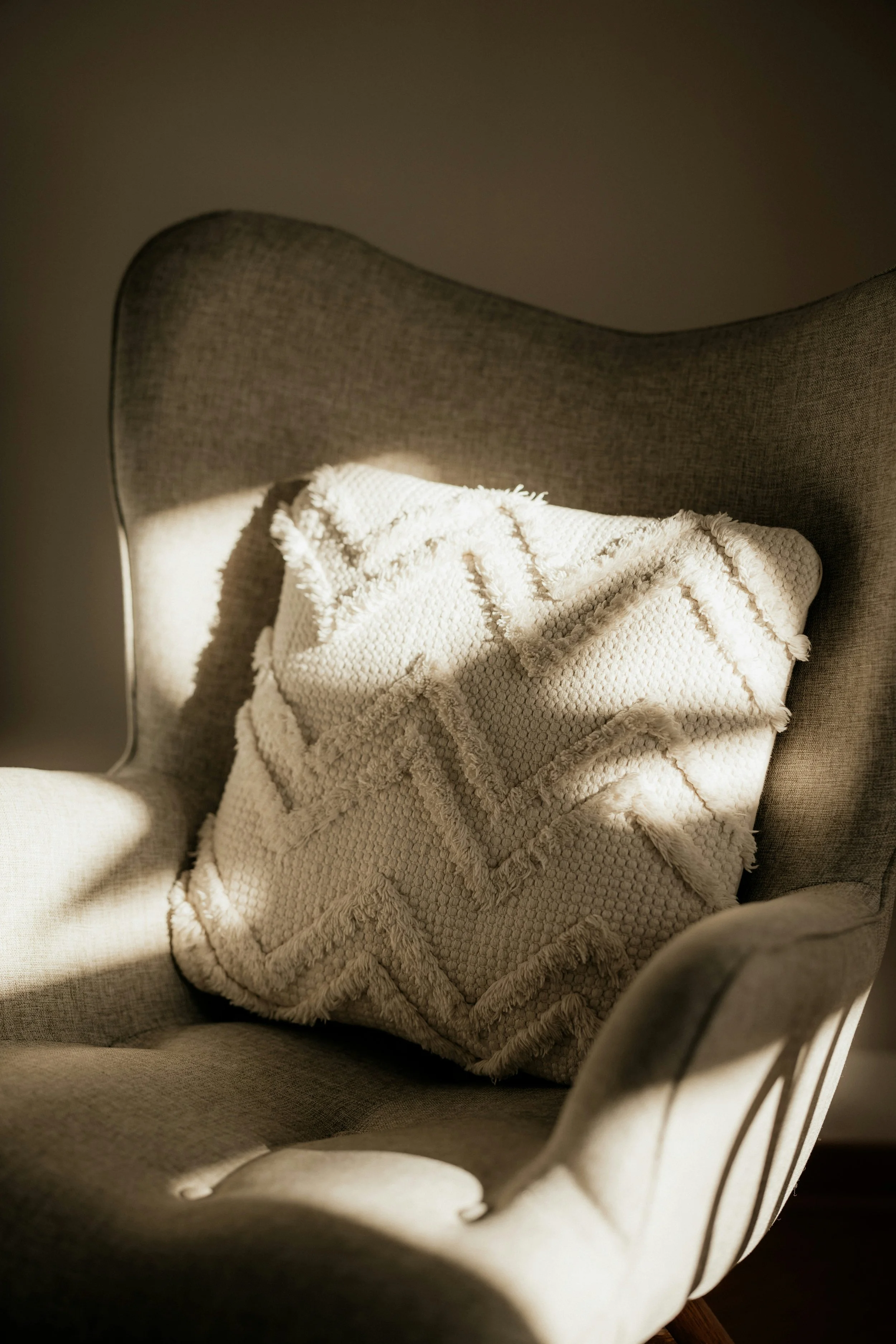 A beige armchair with a textured white pillow featuring zigzag pattern and fringed edges in natural light.