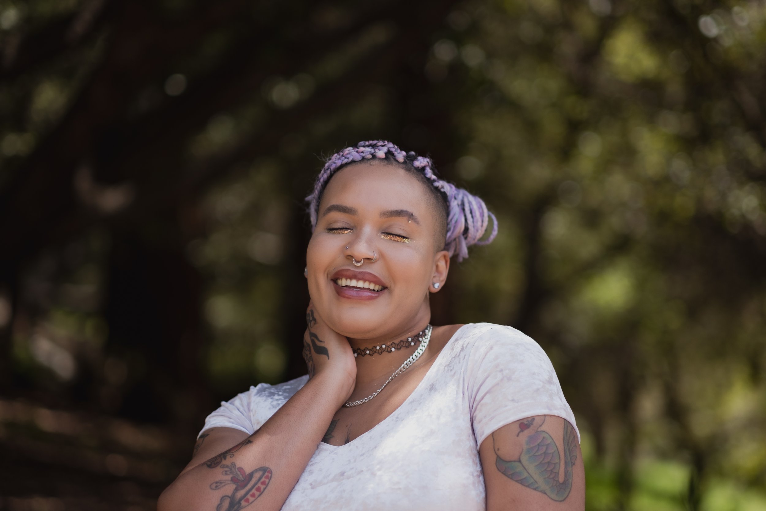 A woman with purple dreadlocks and tattoos on her arms, wearing a white shirt, posing outdoors with trees in the background, smiling with eyes closed.