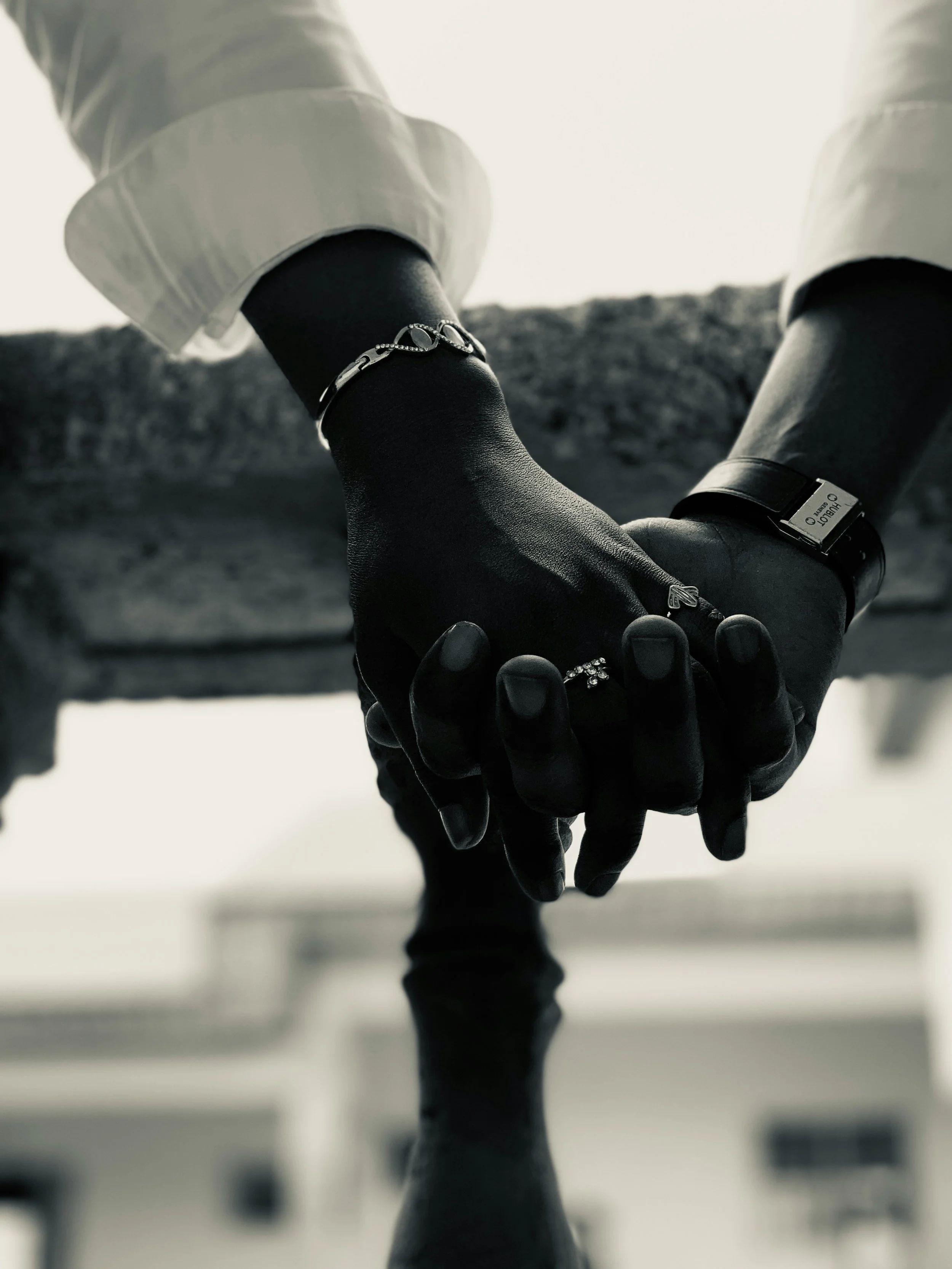 A black and white photo of two hands holding each other, one wearing a watch and ring, the other with a bracelet, against a blurred background.
