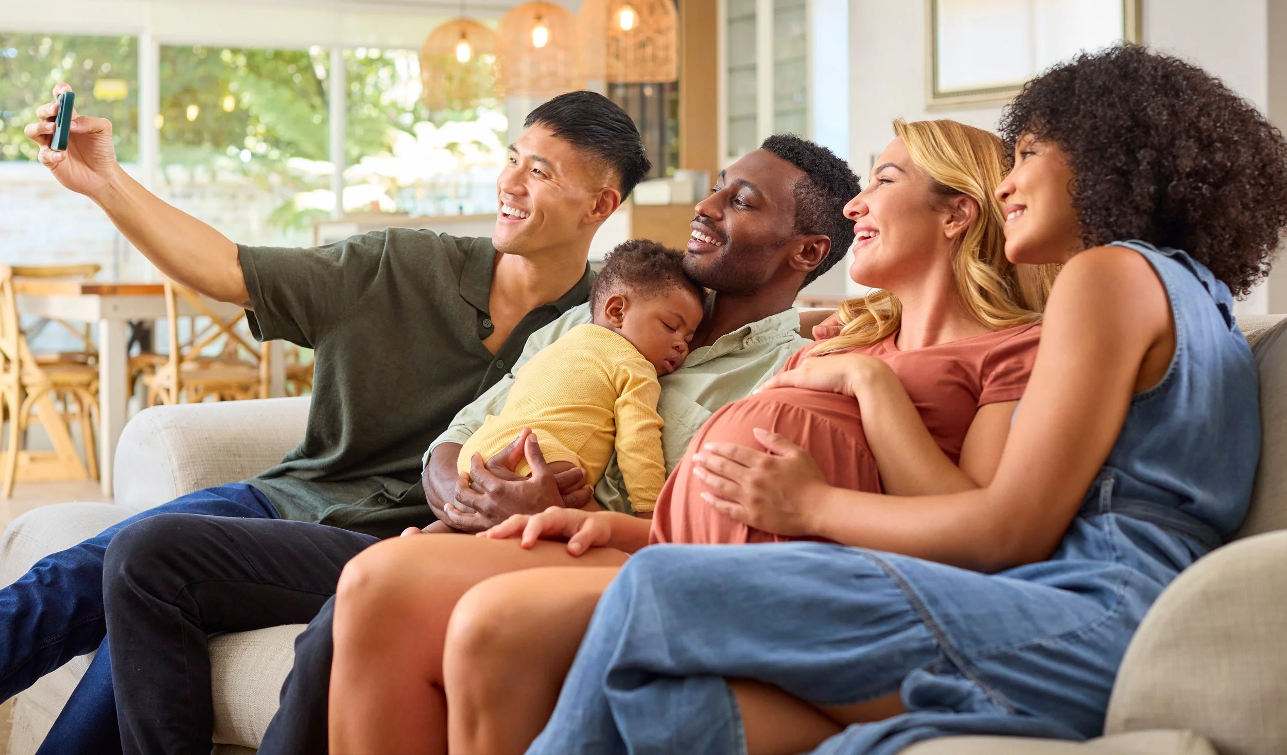 A diverse group of five people, including a pregnant woman, a man holding a sleeping child, and three women, sitting on a couch and taking a selfie together in a cozy, well-lit living room.
