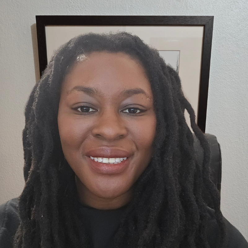 A woman with dark, curly dreadlocks smiling in front of a wall and framed artwork.