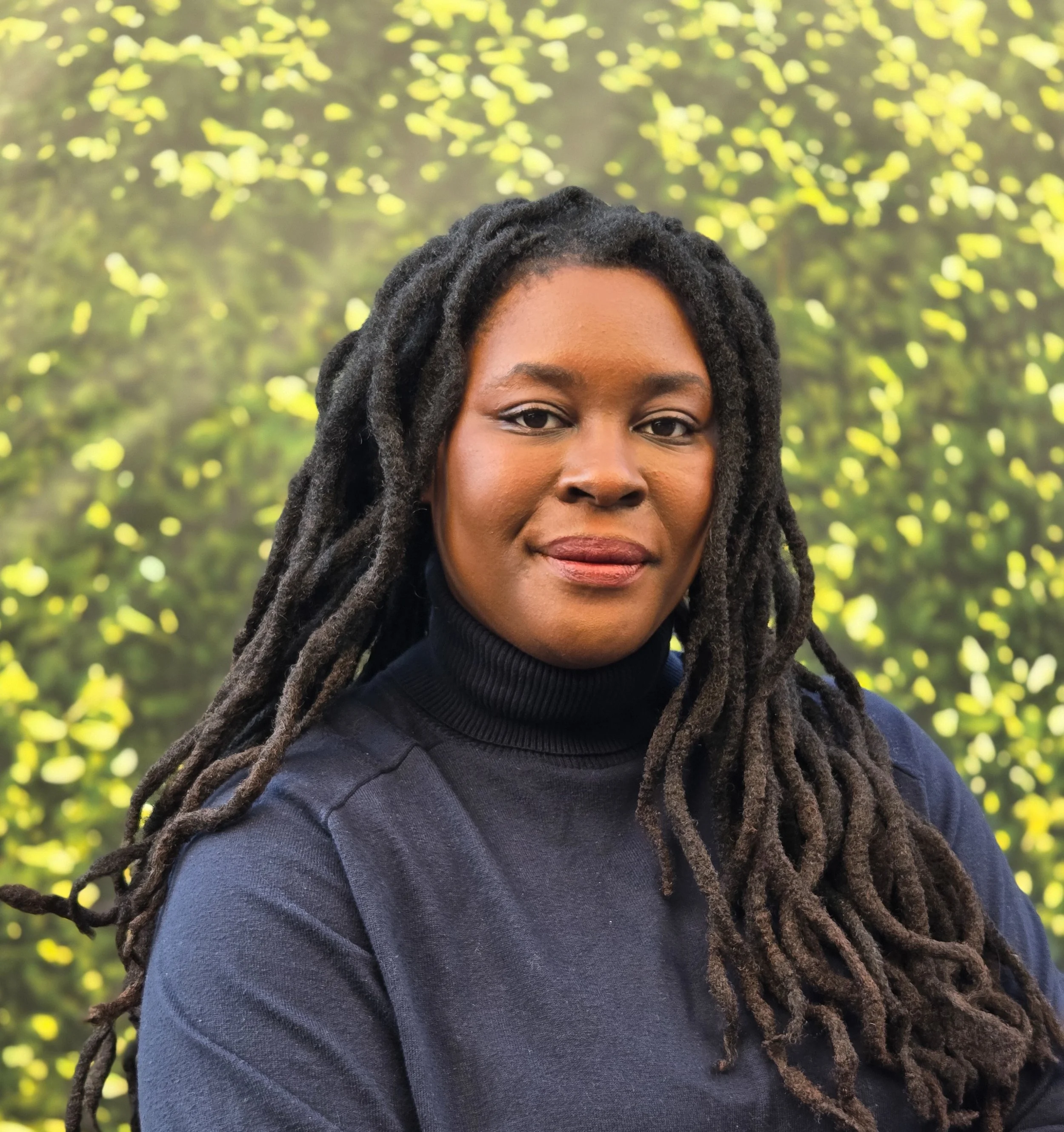 Portrait of a woman with long dreadlocks, wearing a black turtleneck sweater, standing outdoors with blurred green foliage in the background.