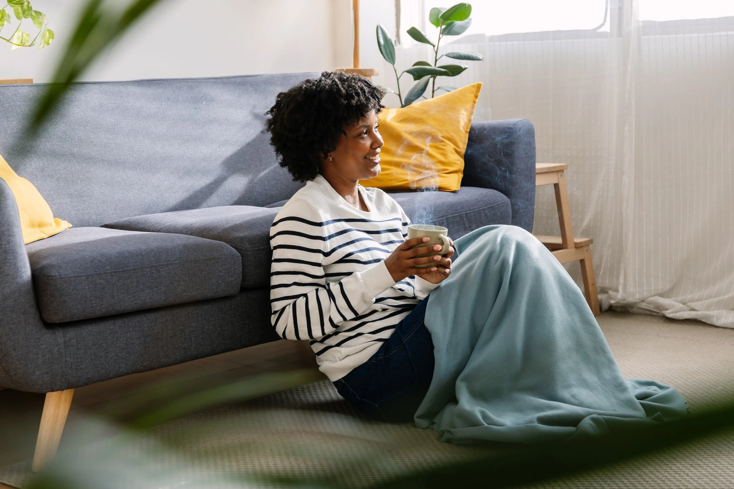 A woman with short curly hair sitting cross-legged on the floor, holding a steaming mug, smiling, next to a gray sofa with yellow pillows, in a well-lit living room with green plants.