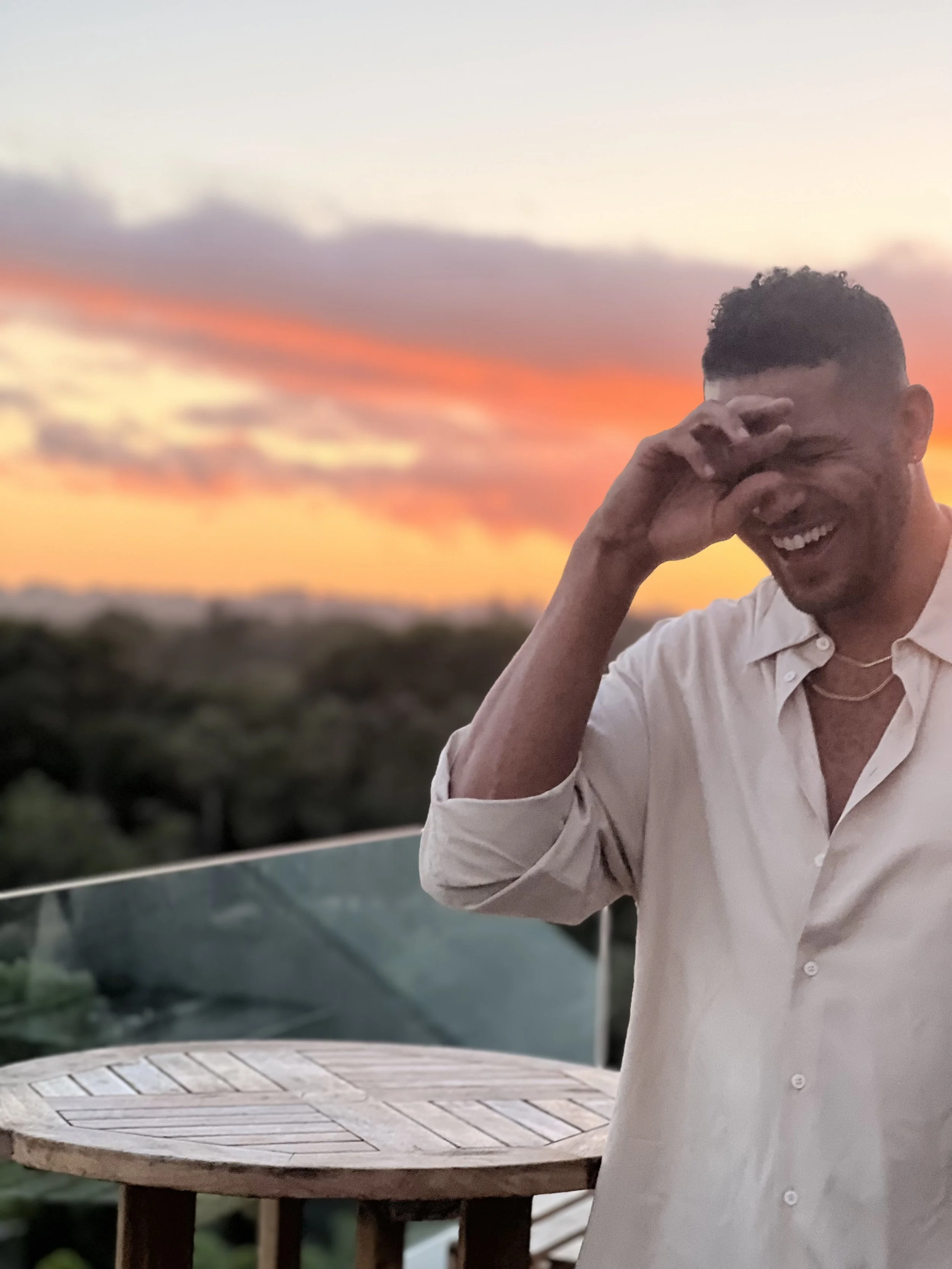 A man named Jeffrey Bowyer-Chapman standing on a balcony overlooking trees and the beach at sunset, He is wearing a white collard shirt with his hand raised up to his head while he is smiling. Middle Frequency.