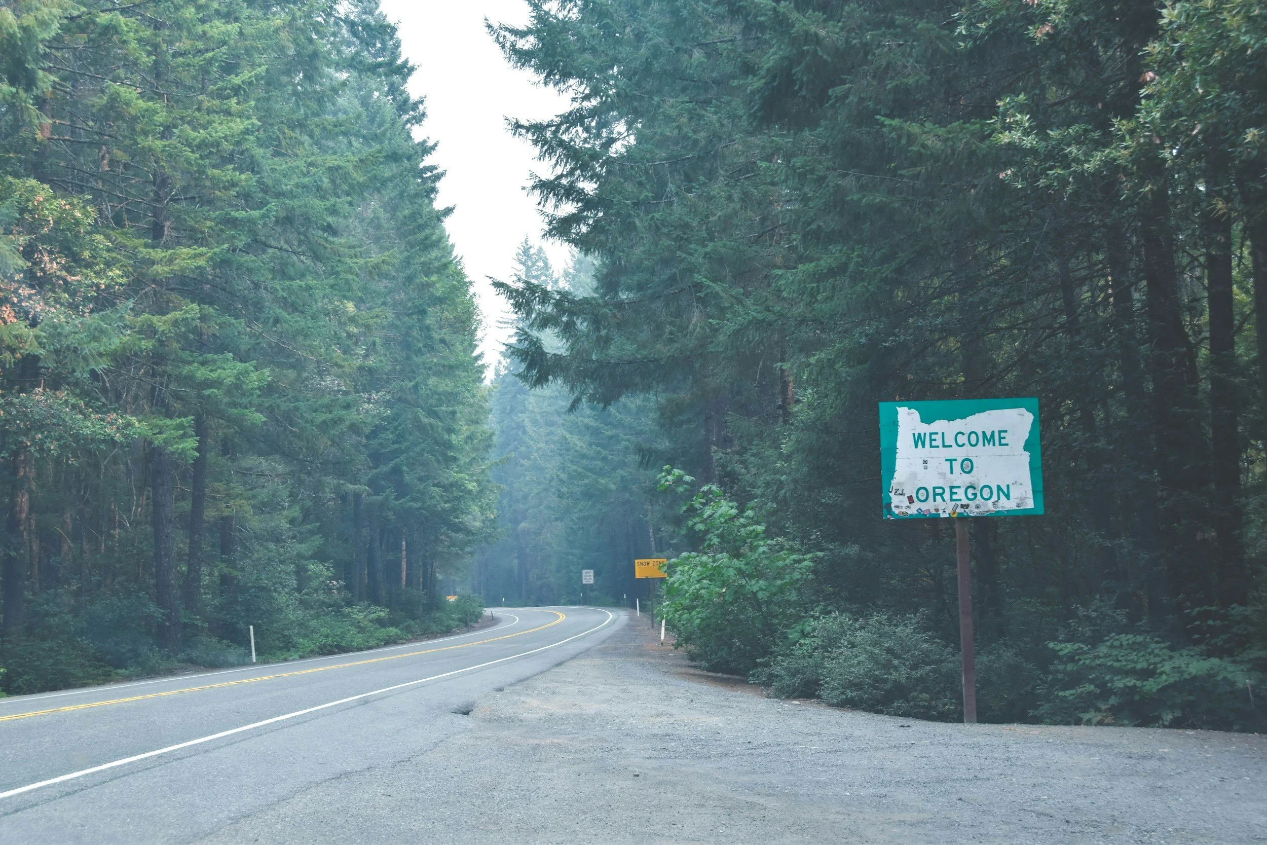 A winding road through a dense forest with tall green trees on both sides. There is a sign that reads 'Welcome to Oregon' along with a yellow warning sign further down the road indicating snow zone.