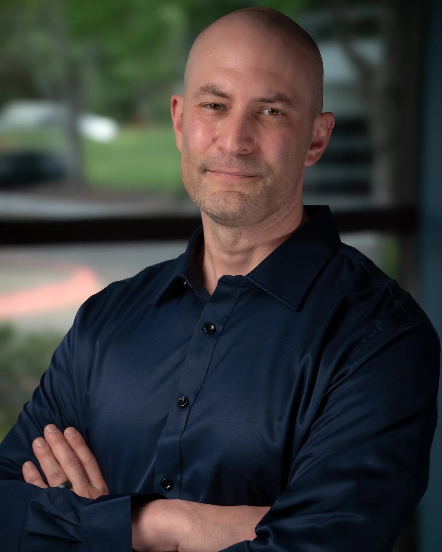 A man with a shaved head, light facial hair, wearing a dark navy blue shirt, standing with his arms crossed inside a building with a blurred outdoor background.