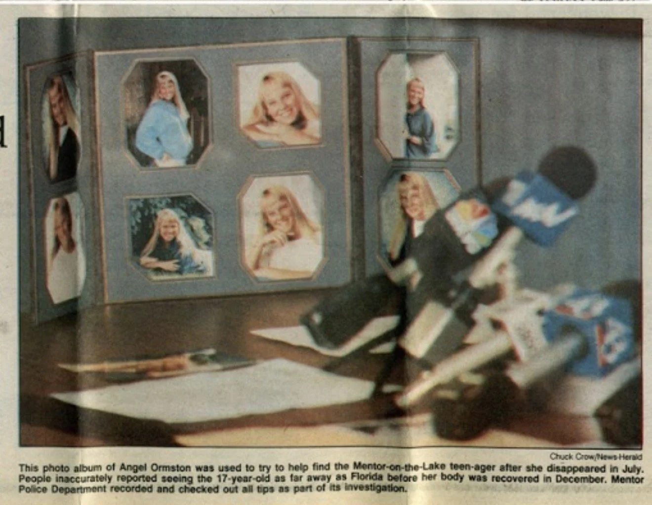 A photo album with multiple pictures of a young girl, Angel Ormiston, displayed on a table. There is a microphone and some papers in front of the album, and a box of tissue nearby. The background is a plain wall.