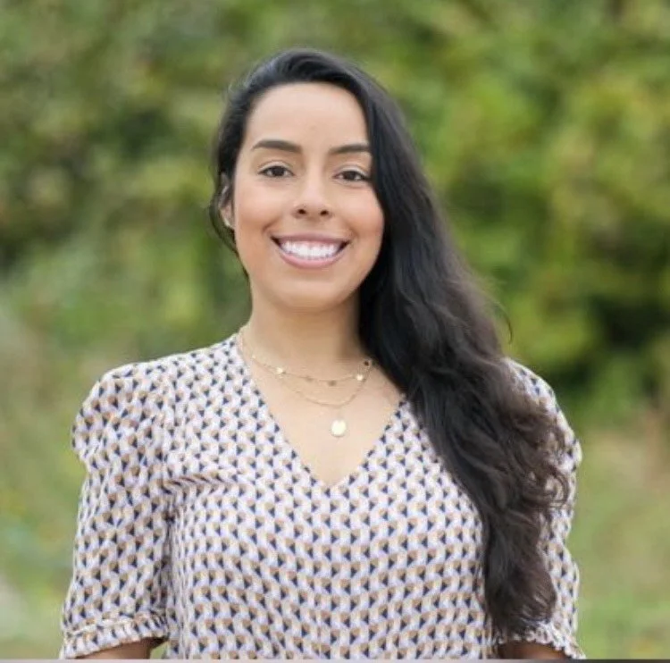 Smiling woman with long dark curly hair wearing a patterned blouse outdoors with green foliage in the background.