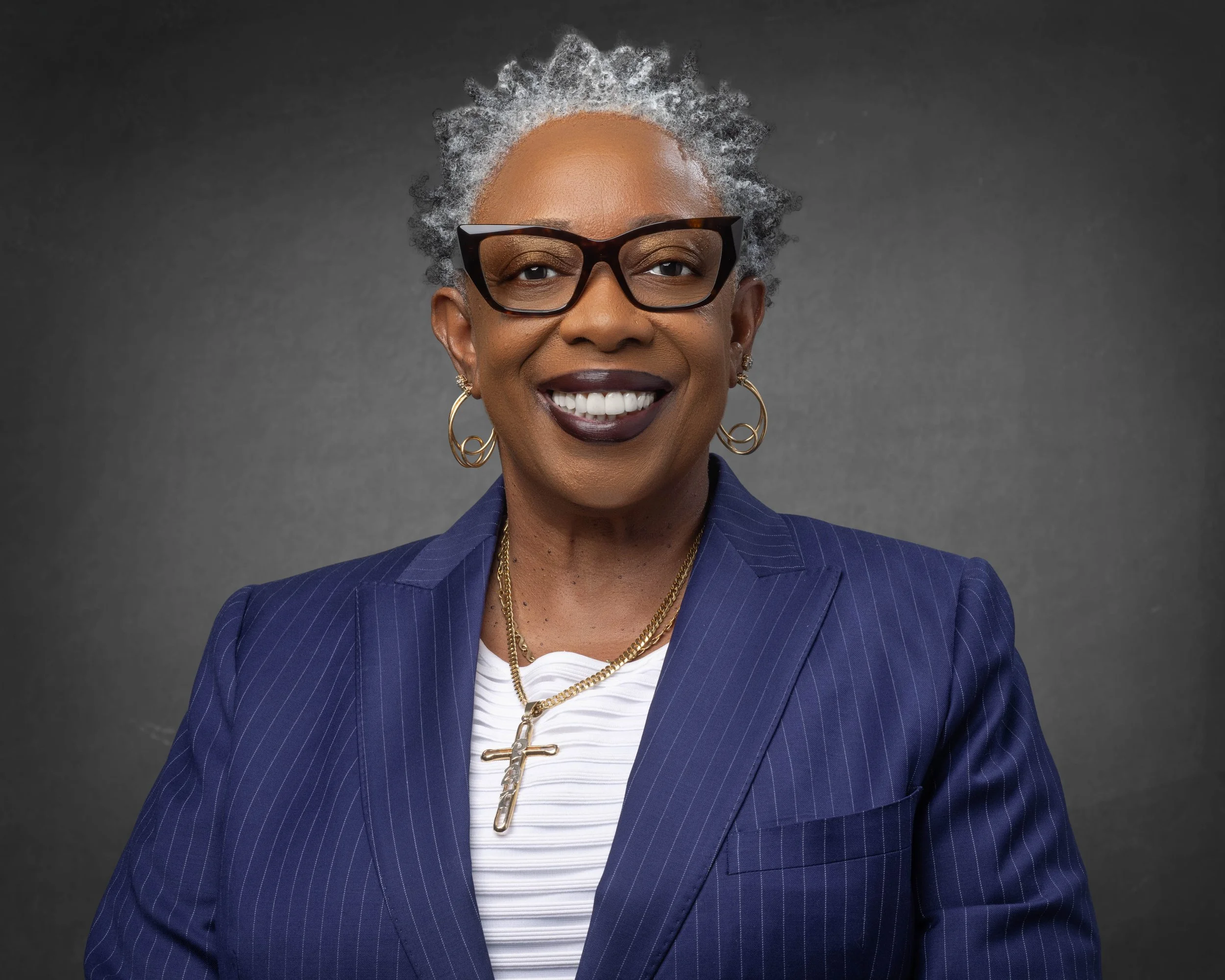 A smiling older African American woman with short gray hair, wearing a navy blue pinstripe blazer, black glasses, gold hoop earrings, and a gold necklace with a cross pendant.