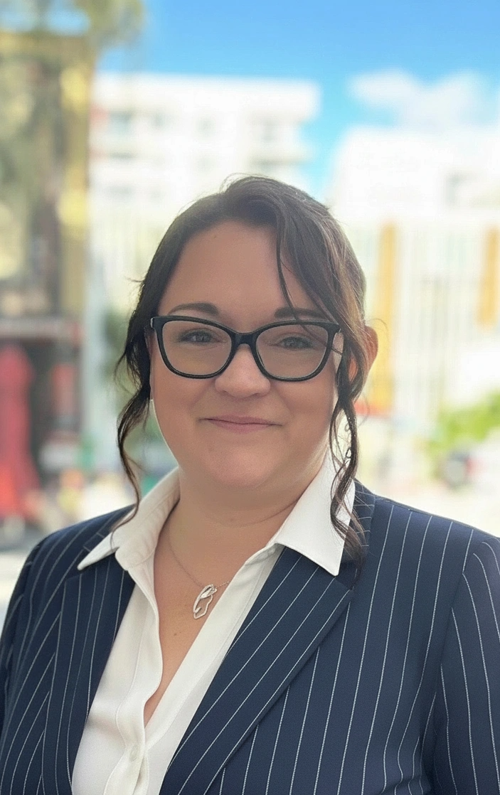 A woman with dark hair, glasses, and a white shirt under a navy pinstripe blazer, standing outdoors with a blurred cityscape background.
