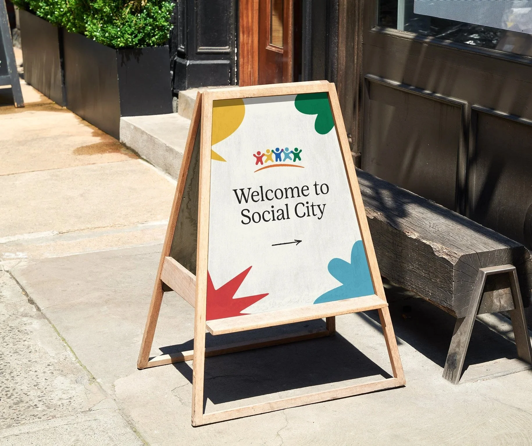 A wooden A-frame sidewalk sign with colorful shapes and text that reads 'Welcome to Social City' and an arrow pointing to the right, placed on a city sidewalk next to a bench and a building entrance.