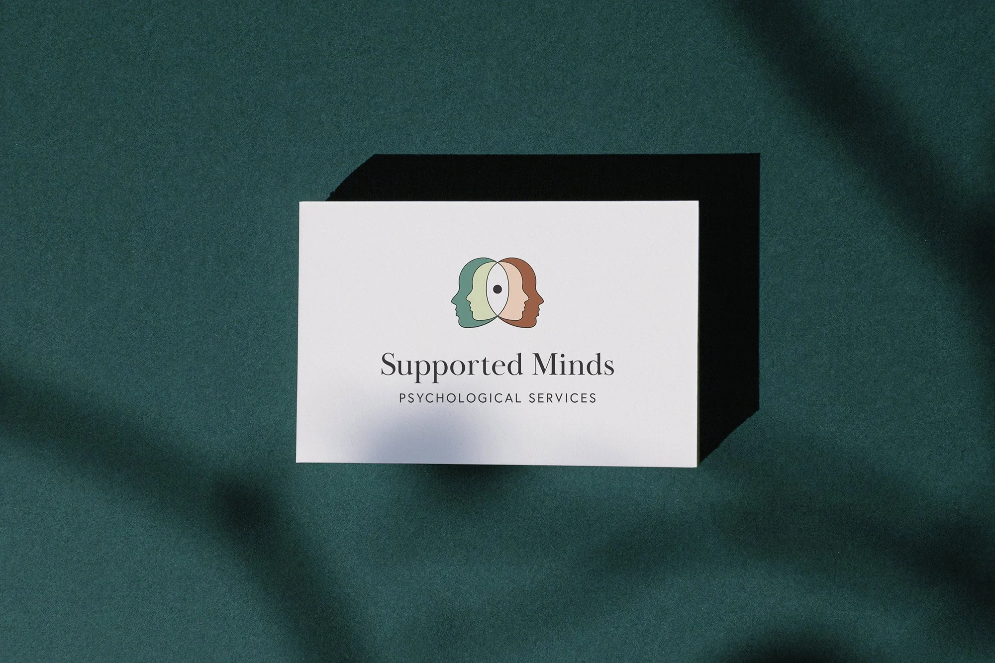 Business card for Supported Minds, a psychological services provider, featuring a logo of three overlapping faces in green, cream, and brown, with a black and white eye in the center.