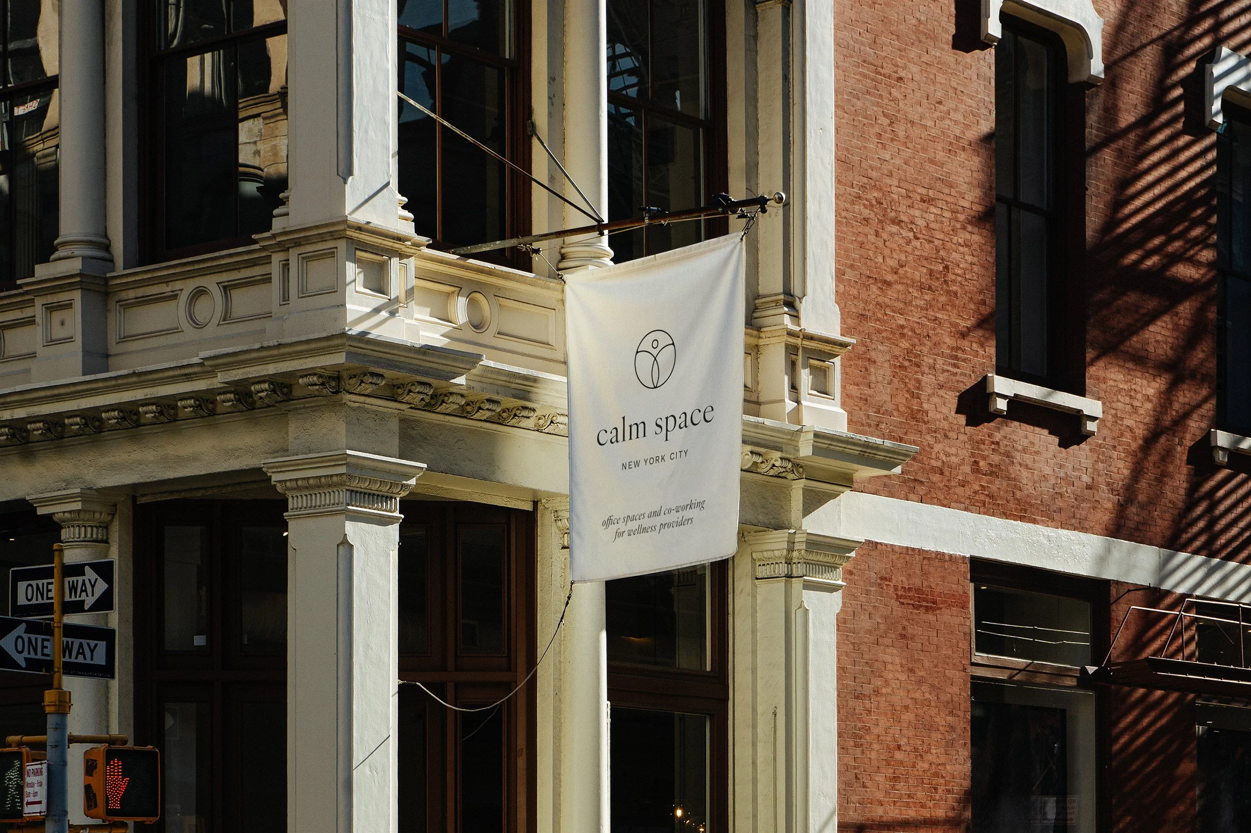 Street view of a building with a sign that reads 'calm space NEW YORK CITY office spaces and co-working for wellness providers'. Shadows from a nearby palm tree are cast on the brick wall.