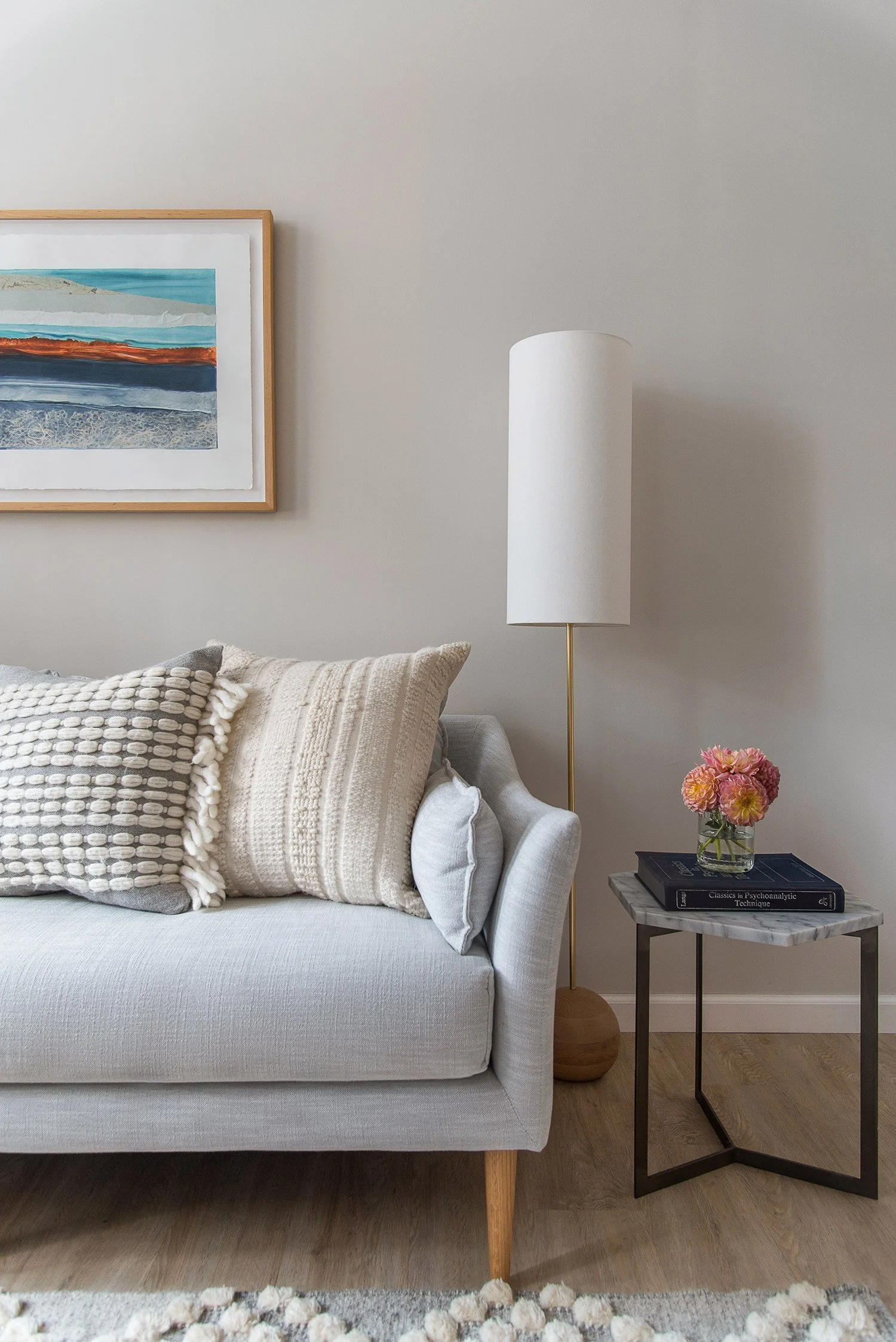 Living room with a light-colored sofa decorated with textured pillows. A side table with a stack of books and a vase with pink flowers. A tall white floor lamp next to the sofa. A framed colorful landscape painting on the wall.