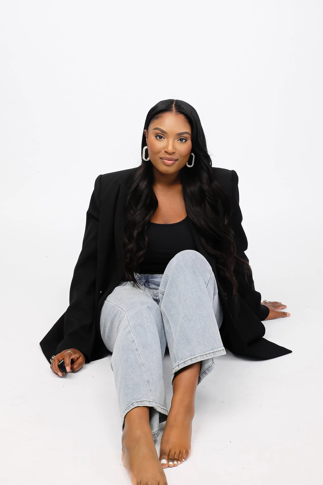 A woman with long dark hair, wearing hoop earrings, a black top, a black blazer, and light-wash jeans, sitting barefoot on a white background.