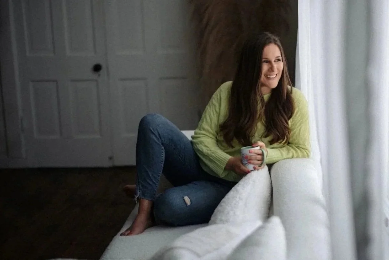 Emma with long brown hair, wearing a light green sweater and ripped jeans, sitting on a white couch by a window, holding a mug, smiling while looking outside.