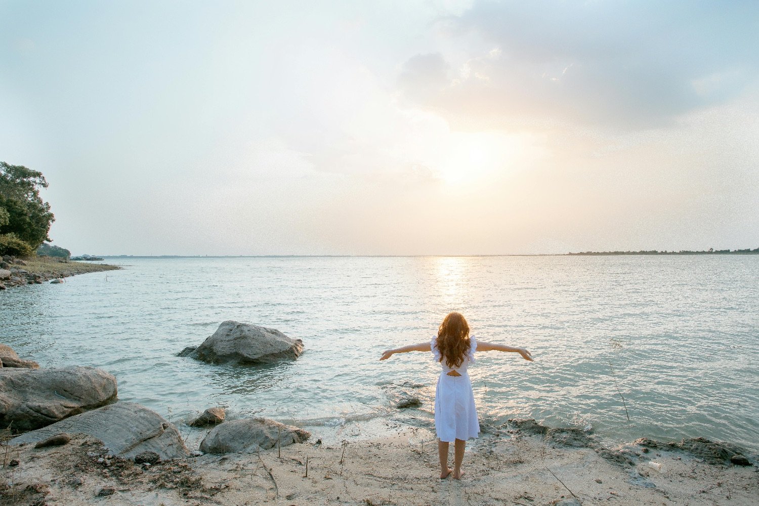 A woman with long hair in a white dress stands barefoot on a sandy beach with rocks, facing water with her arms outstretched, during sunset or sunrise.