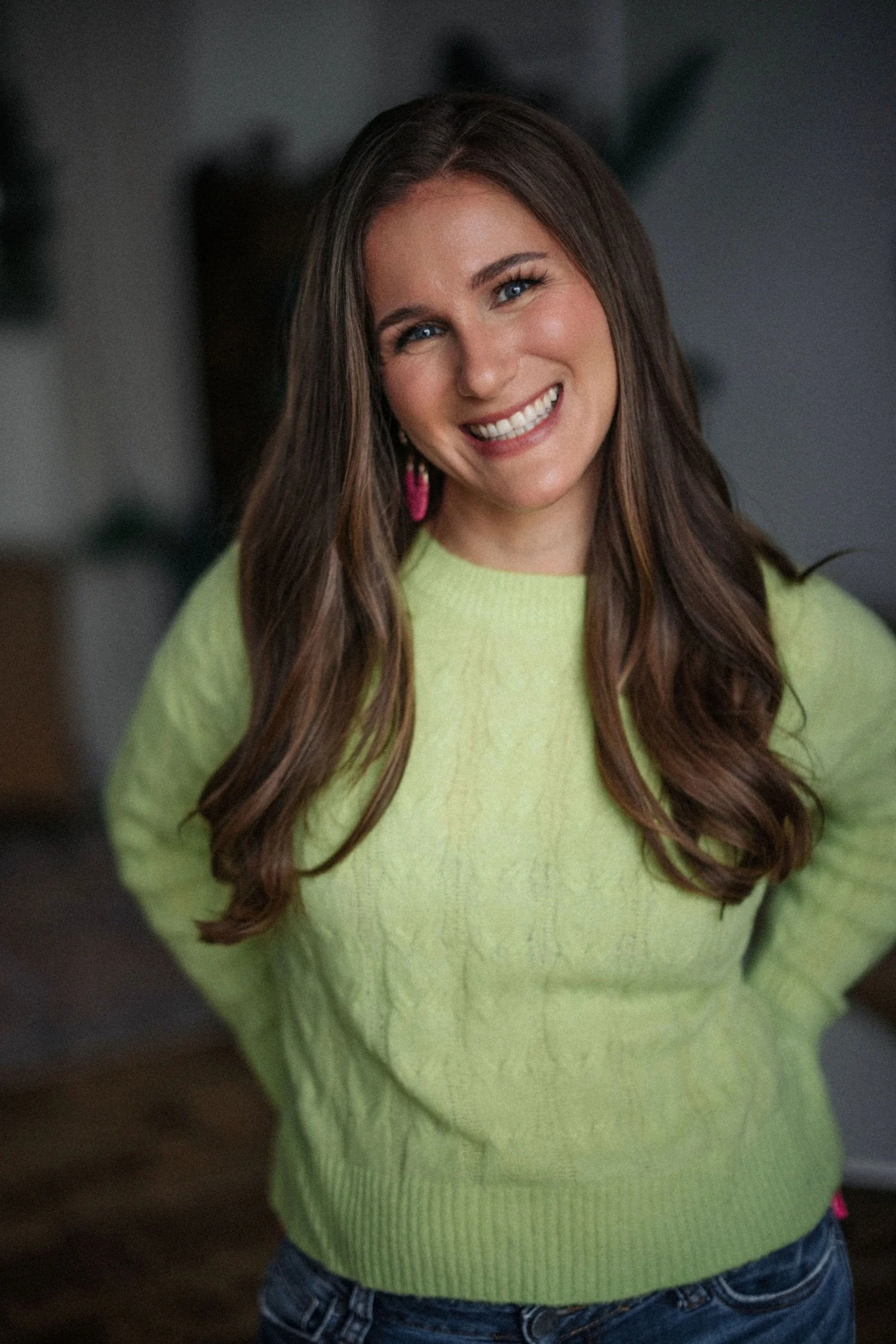 Emma with long brown hair, blue eyes, and a bright smile wearing a lime-green sweater and pink earrings, standing indoors.