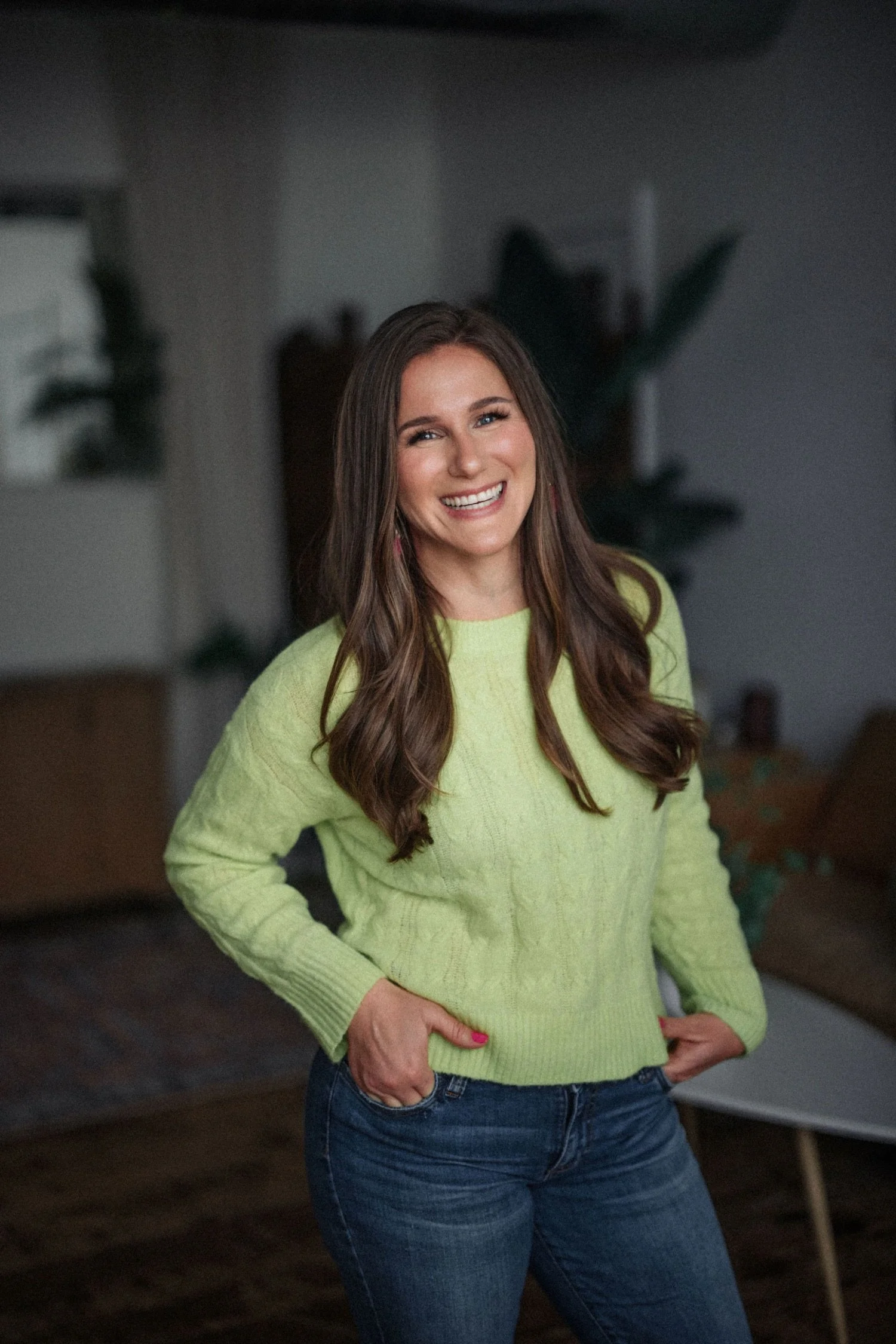 A smiling Emma with long brown hair wearing a lime green sweater and jeans, standing indoors with hands in her pockets.