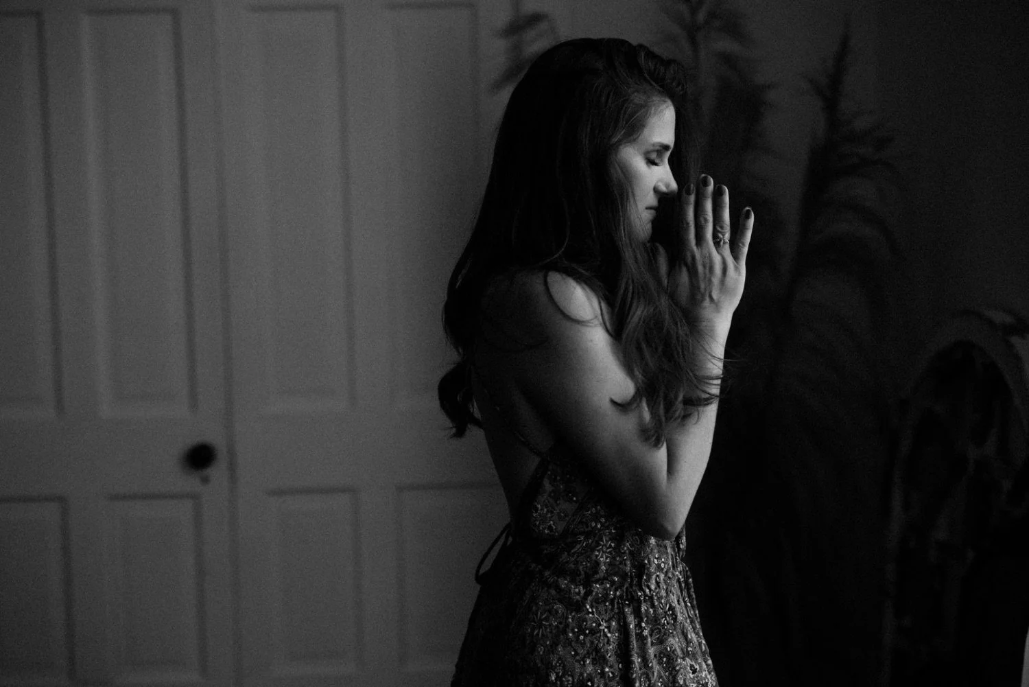 Emma with long wavy hair standing indoors with her eyes closed, hands pressed together in prayer or meditation, wearing a patterned dress.