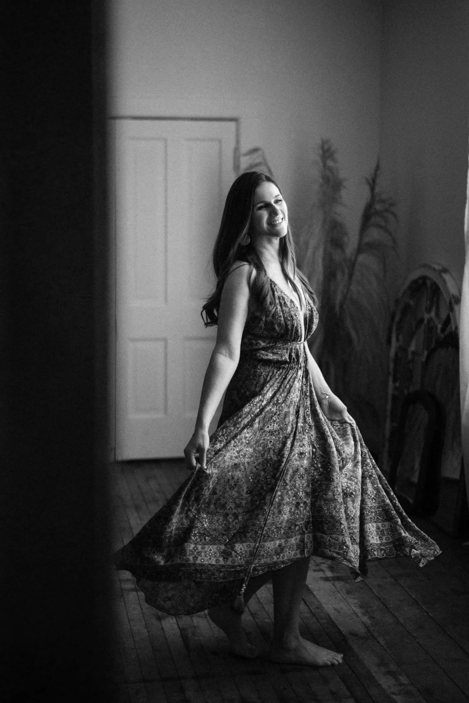 Emma with long hair wearing a patterned dress, smiling while twirling barefoot on a wooden floor in a softly lit room, with plants and a closed door in the background.