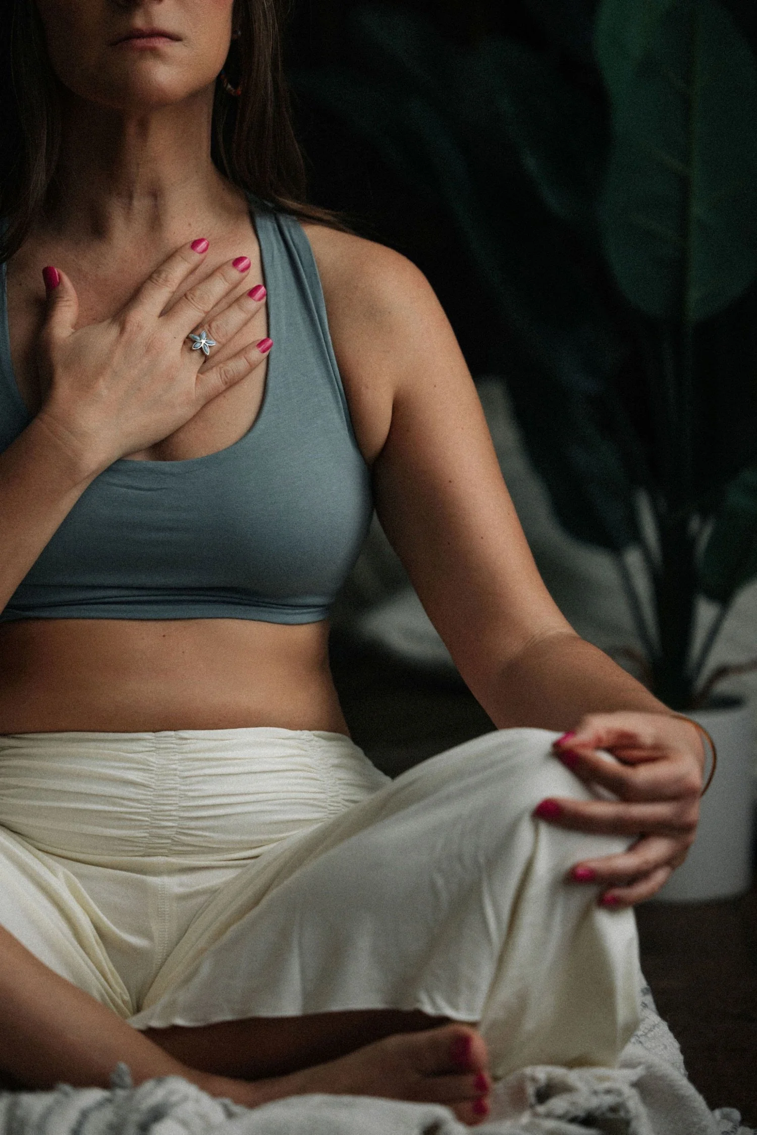 Emma in a blue sports bra and cream-colored pants sits cross-legged, resting her hand on her chest and her other hand on her knee. She has pink painted nails and is wearing a flower-shaped ring. The background contains dark leafy plants.