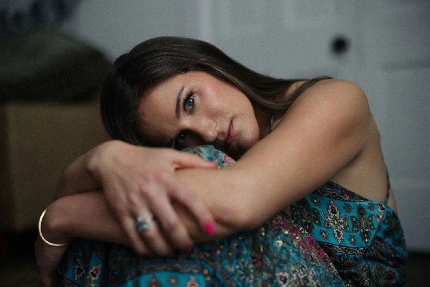 Emma who has long brown hair and blue eyes rests her head on her crossed arms, wearing a colorful patterned top, looking directly at the camera.