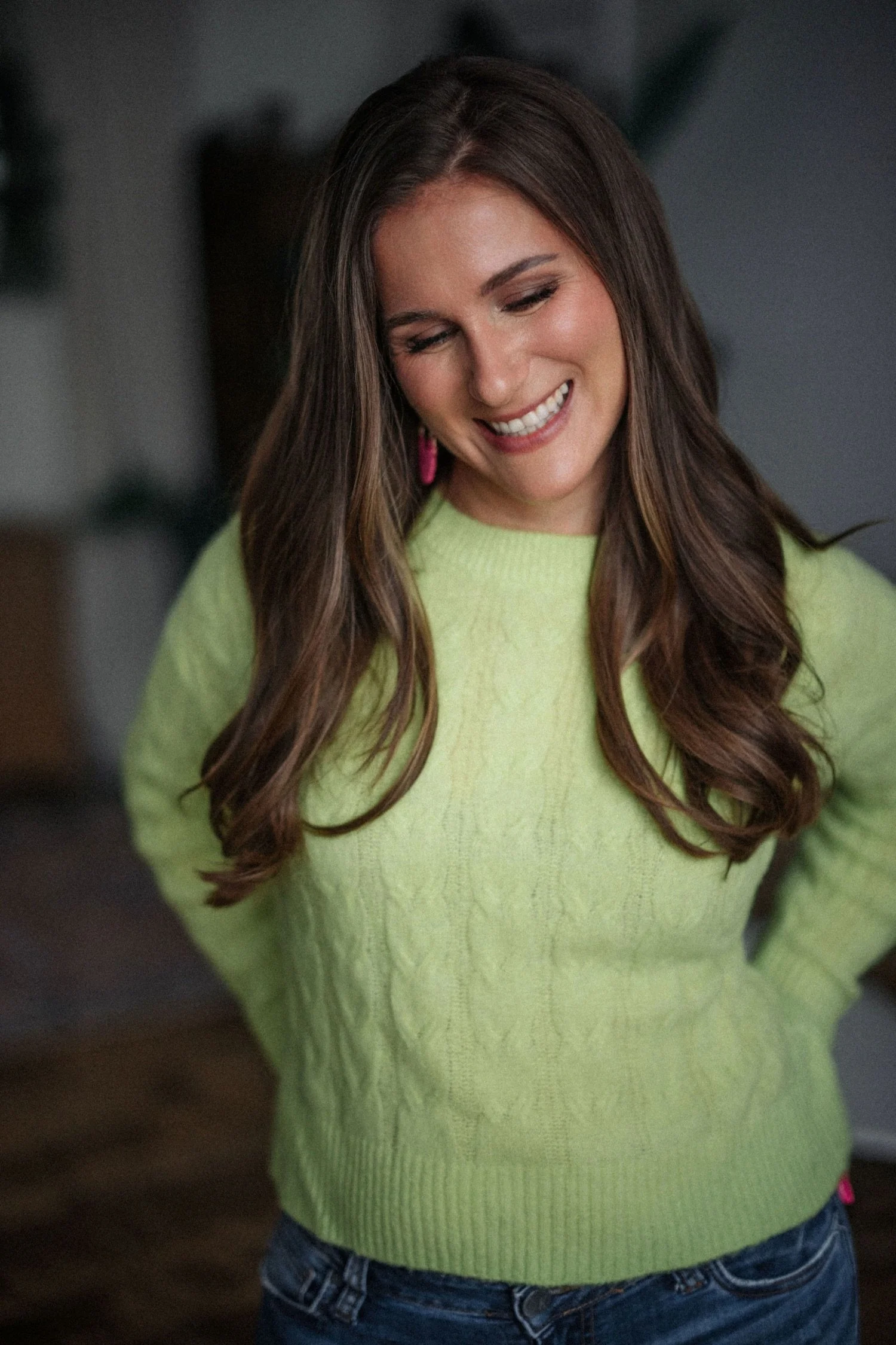 Emma, who has long brown hair, smiling with eyes closed, wearing a lime green sweater and blue jeans.