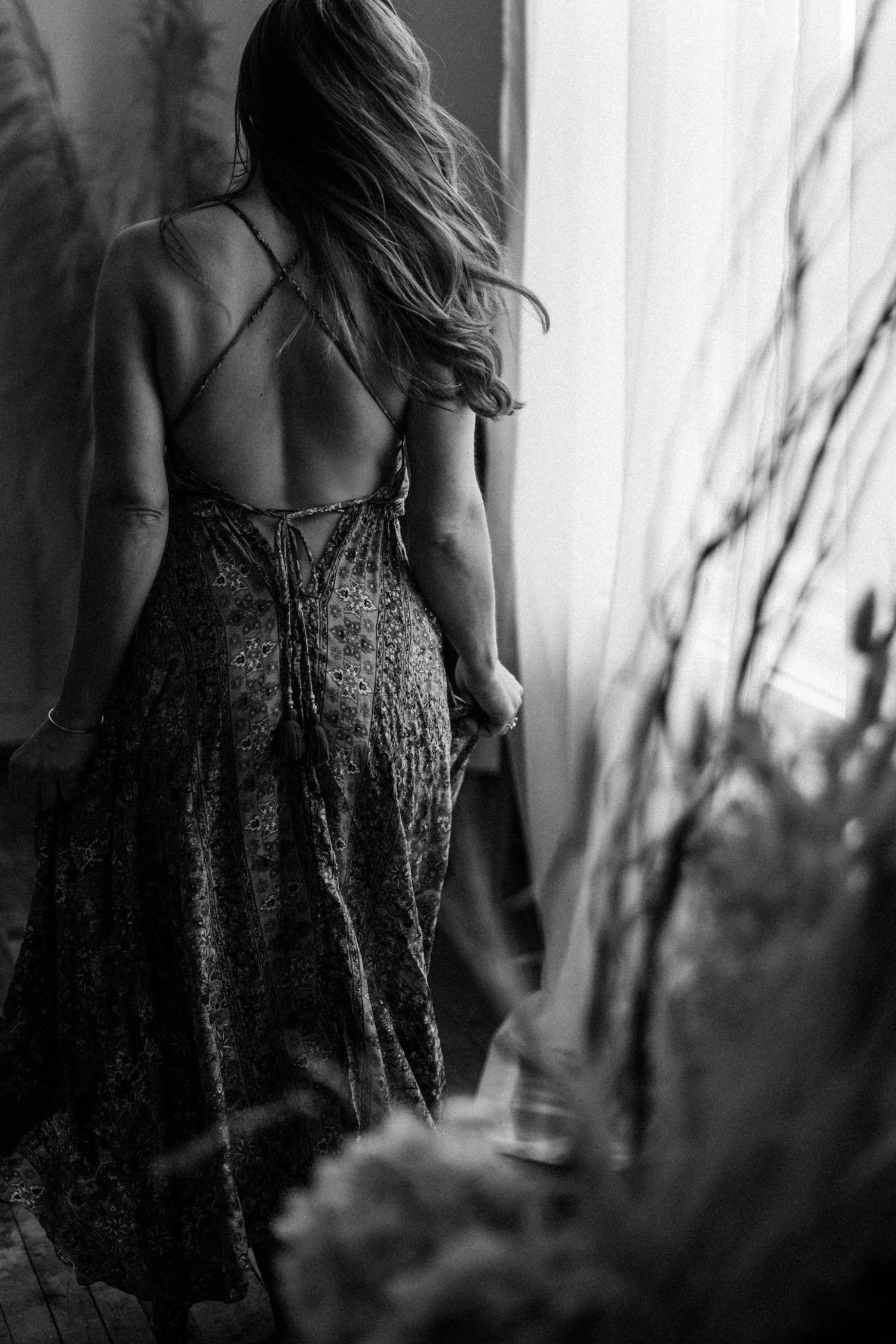 Emma with long hair, dressed in a patterned backless dress, stands near a window, with her back facing the camera in a black and white photo.
