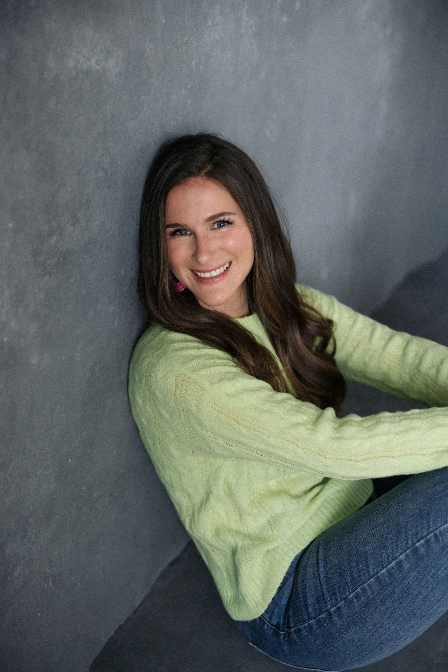 Emma with long brown hair, wearing a light green sweater and blue jeans, sitting on the floor and smiling at the camera against a gray wall.