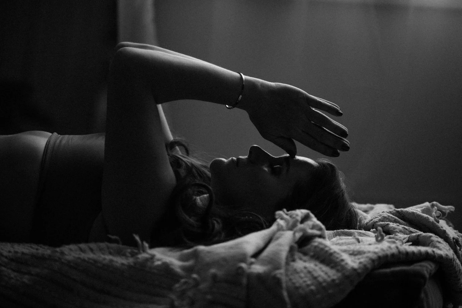 A black-and-white photograph of Emma lying on her back on a bed with a textured blanket. She is resting her head on a pillow, with her eyes closed, and her hands are pressed together raised above her face with her fingers  touching her forehead