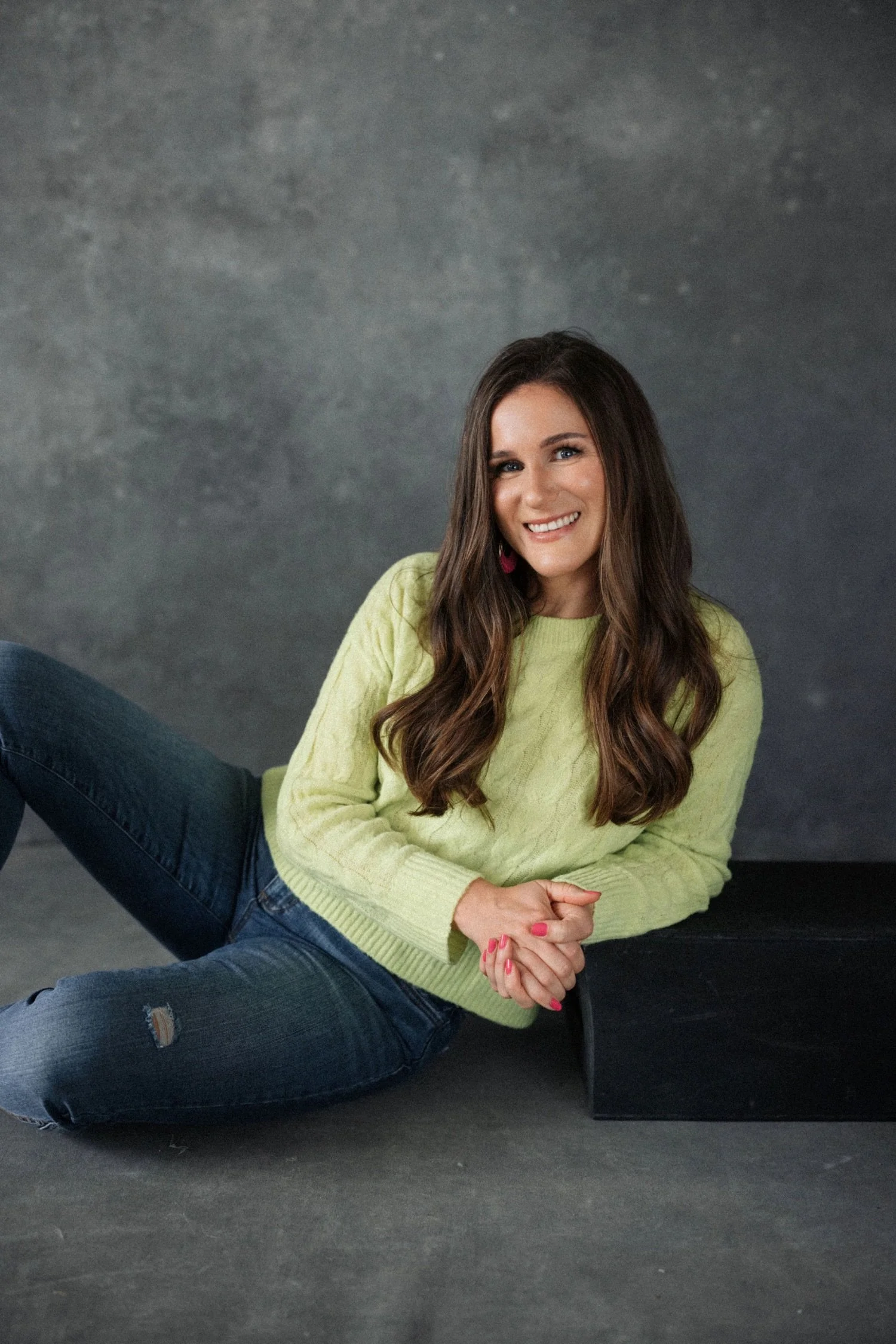 Emma with long brown hair is lying on the floor, smiling, wearing a light green sweater and dark jeans, with a dark gray background.