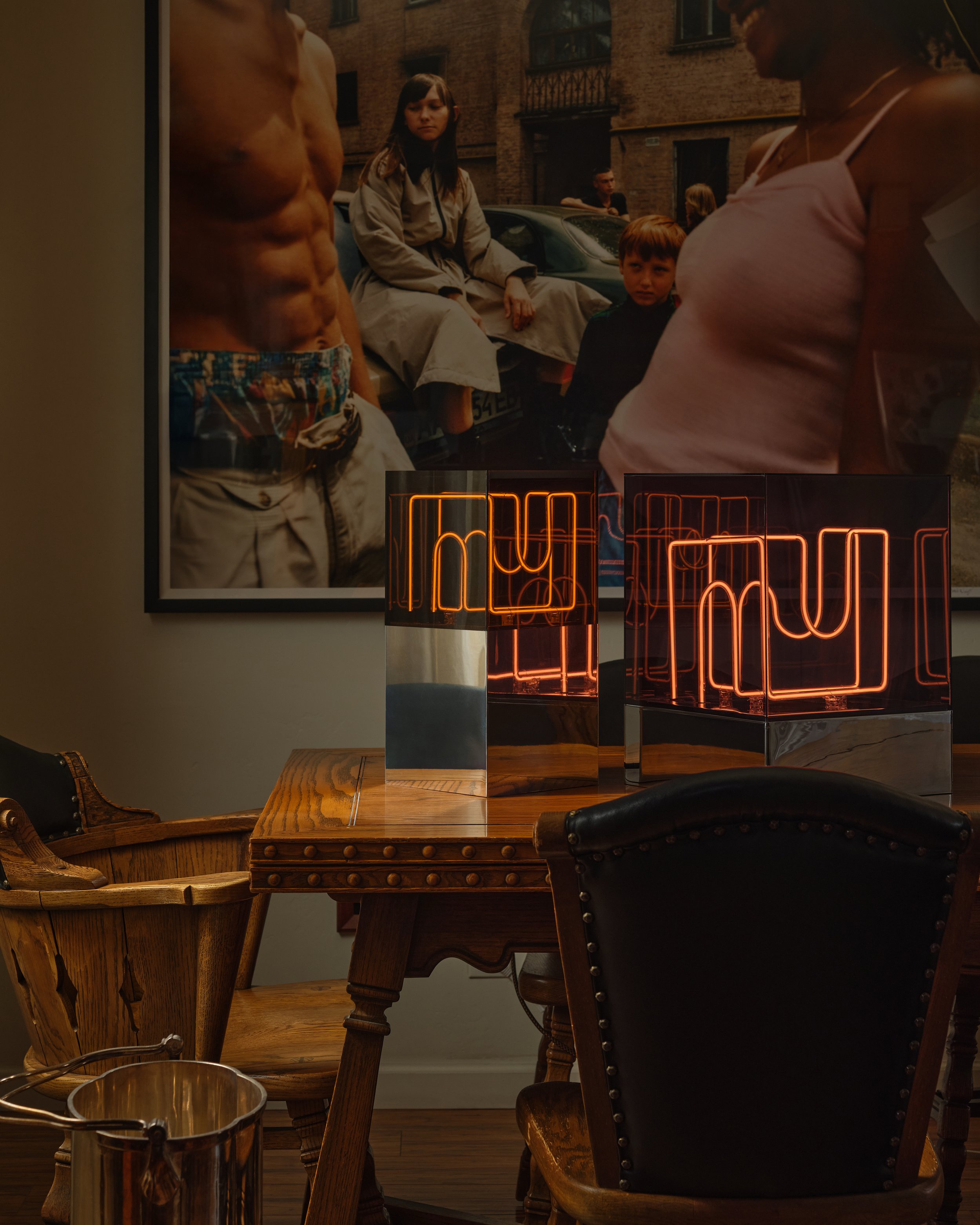Photo of a decorated room with a large framed photo on the wall, showing people on a street. On a wooden table, a neon sign spells 'muh' in orange lights, and a black leather chair with nailhead trim is partially visible.