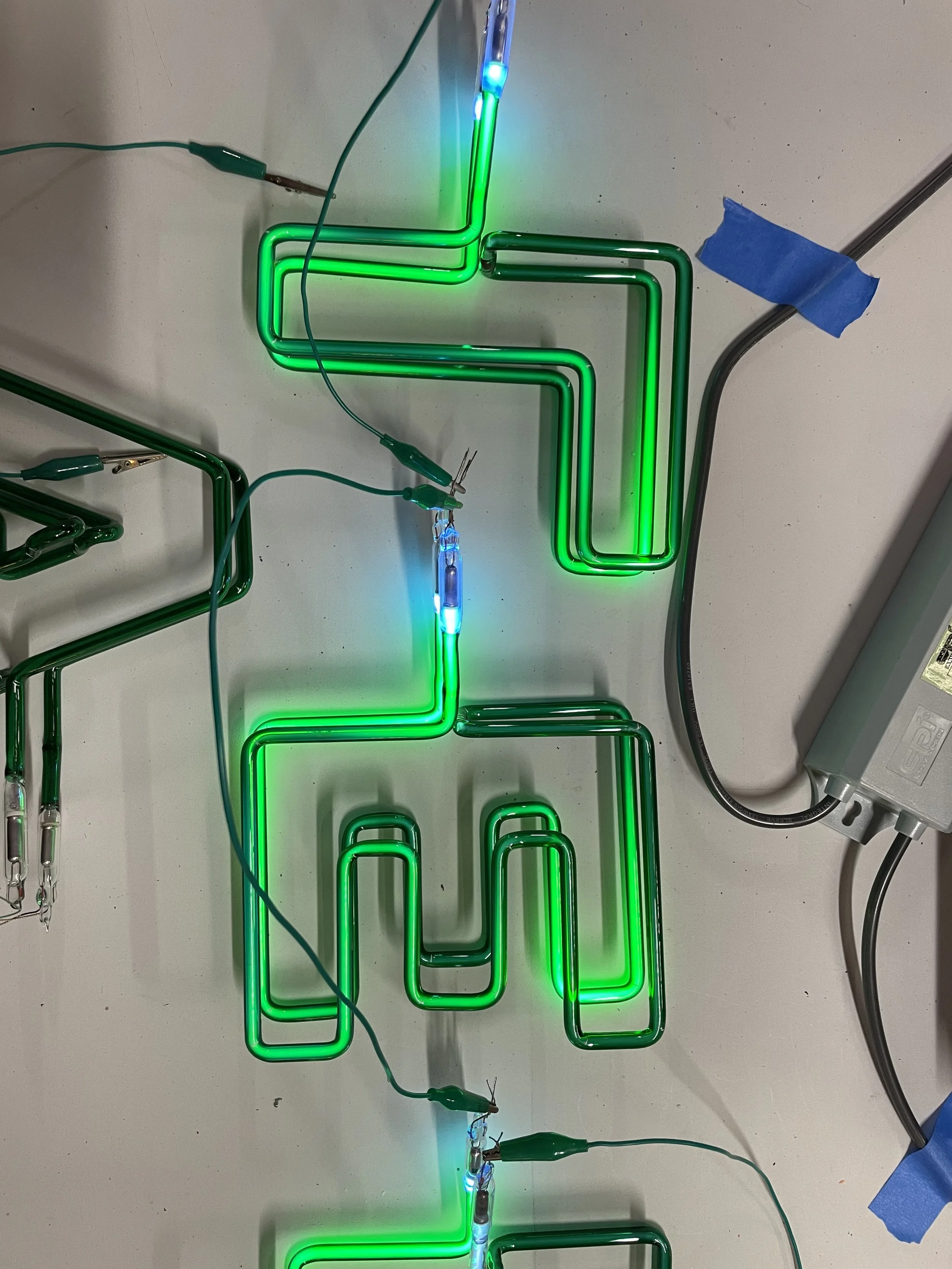 Close-up of a neon green and blue L-shaped electronic circuit on a white surface, with wires and electrical connections, part of a technical experiment or demonstration.