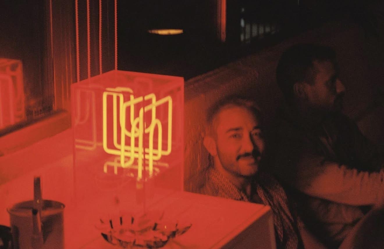 A neon sign with red Chinese characters, a man with a mustache and short hair smiling, and another person with short hair looking away, sitting on a bench or similar surface in a dimly lit setting.