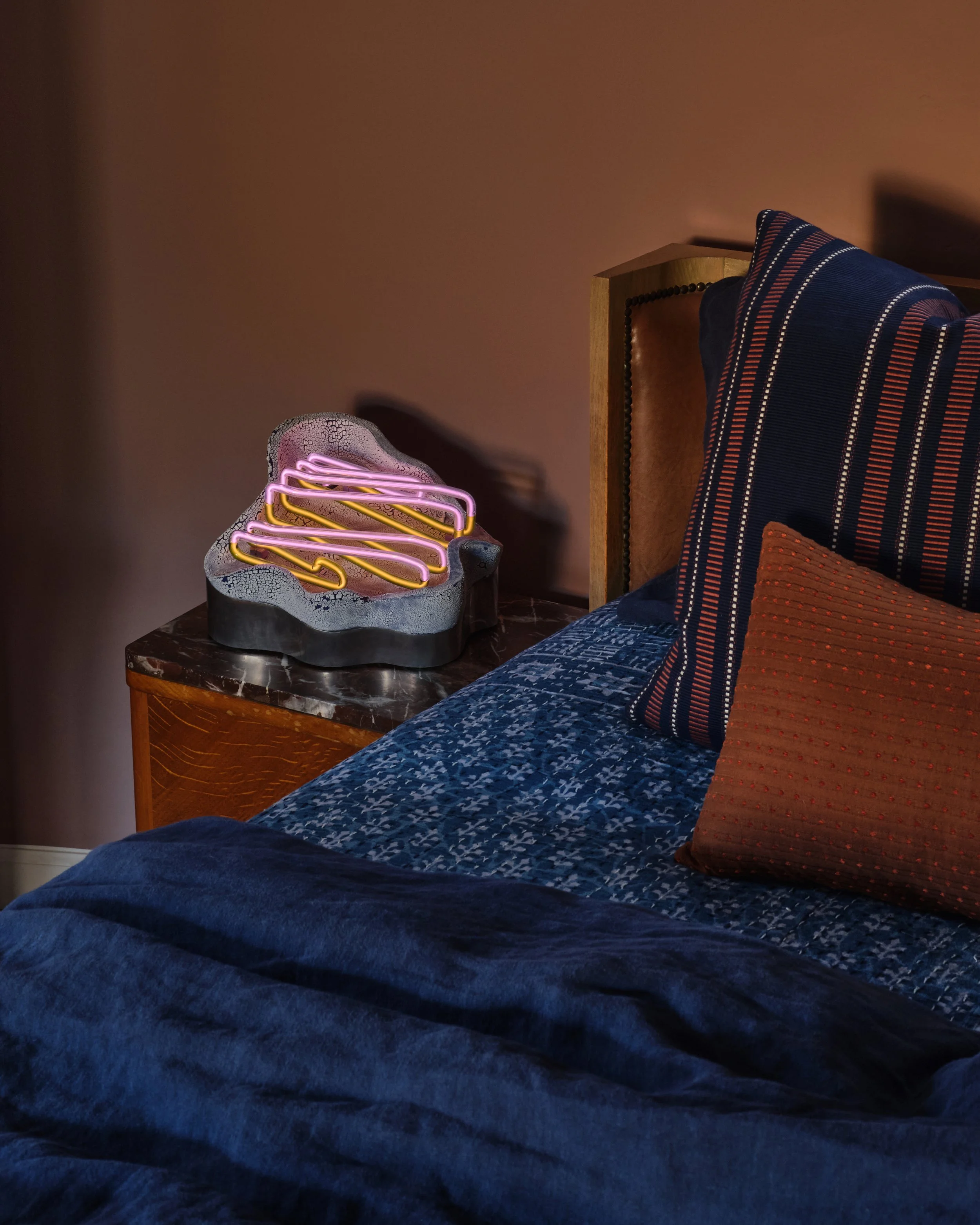 A bedroom with a blue bedspread, striped pillows, and an eclectic bedside table featuring a bowl with pink and yellow neon lights shaped like a cloud.