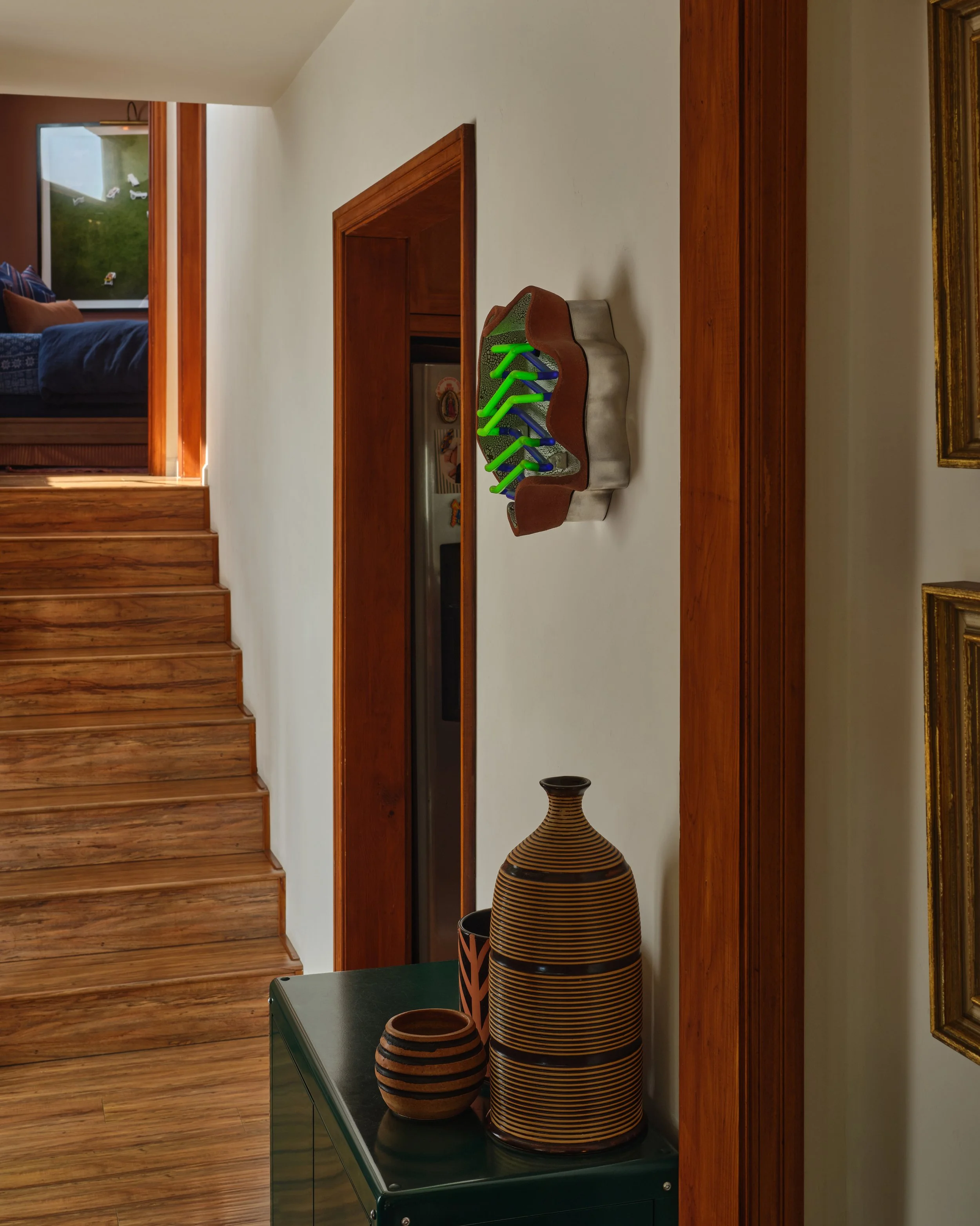 Interior view of a home with wooden stairs, a wall-mounted decorative shoe display with colorful shoes, and a table with striped pottery.