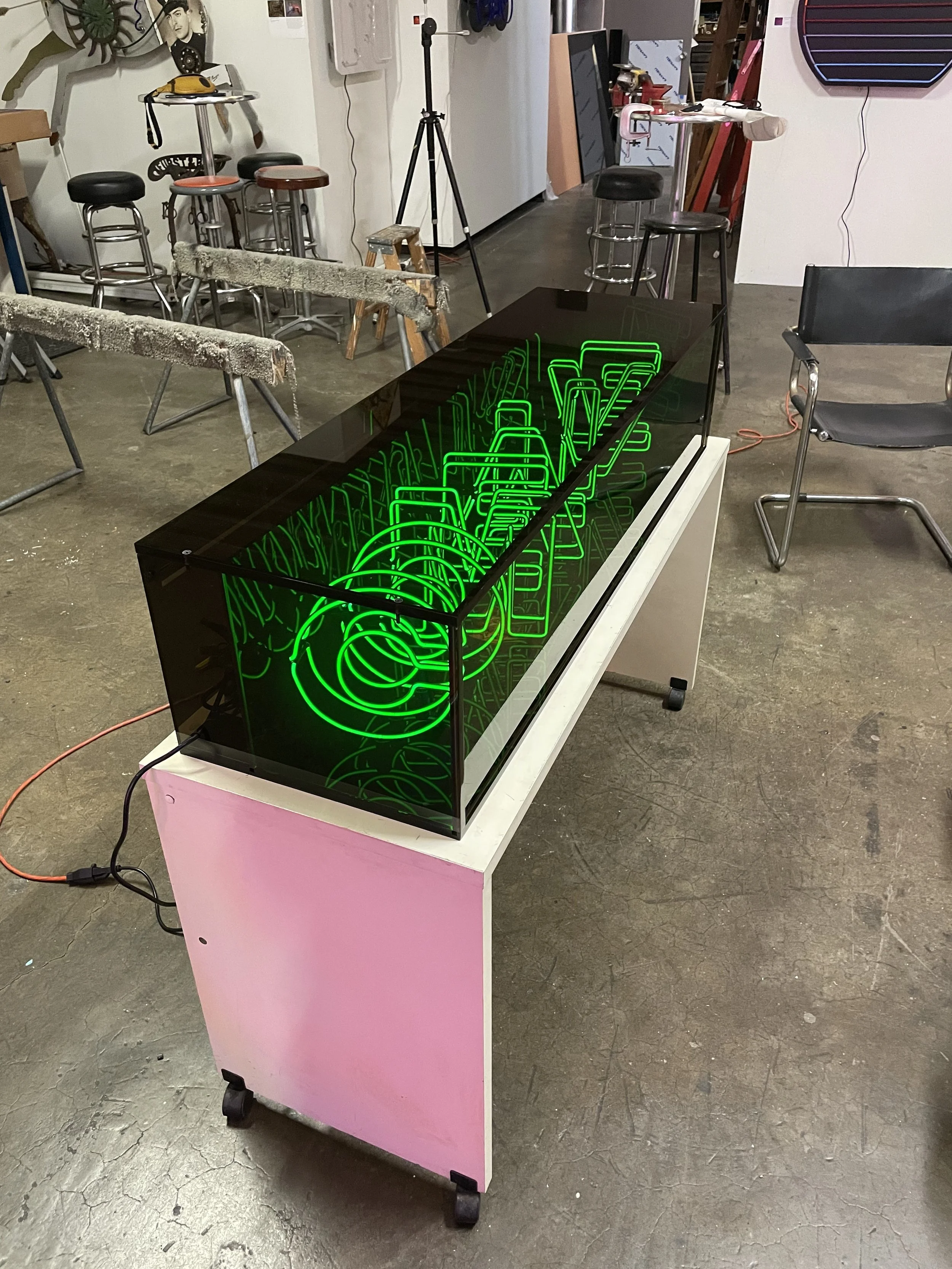 A glass display case with c-shaped green neon light tubes inside, placed on a pink and white cart in a workshop or art studio, with tools, chairs, and furniture in the background.