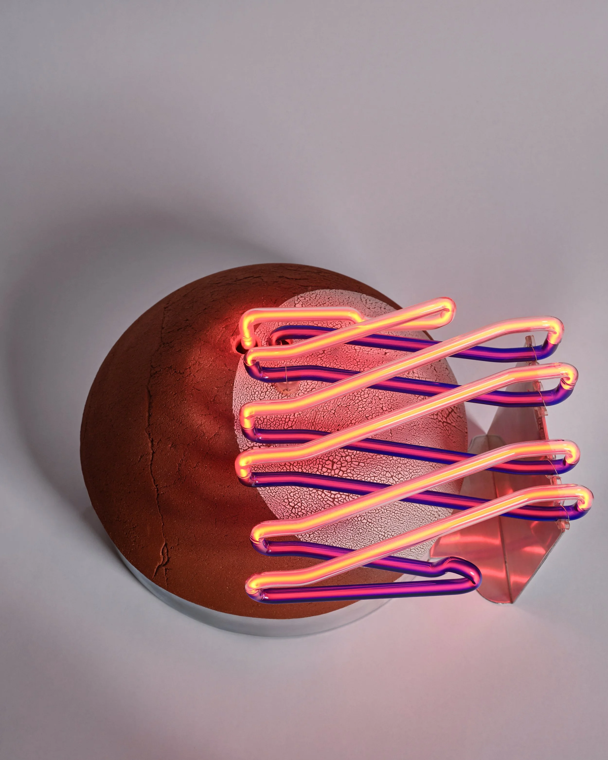 A neon art installation with pink, orange, and purple glowing tubes over a cracked, textured, round brown surface.