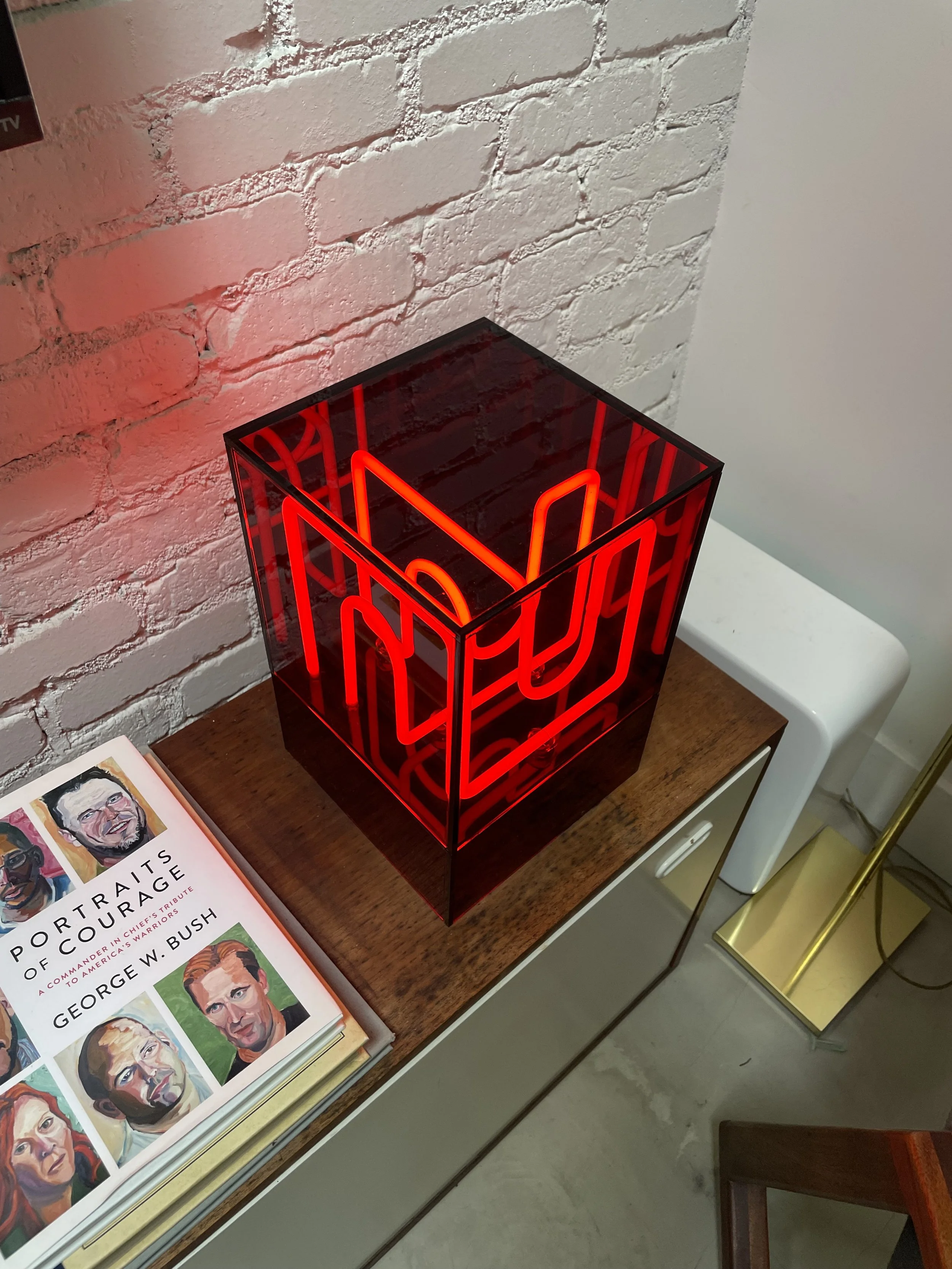 Neon red sign inside a transparent cube on a wooden table, next to a book about George W. Bush, with a white brick wall in the background.