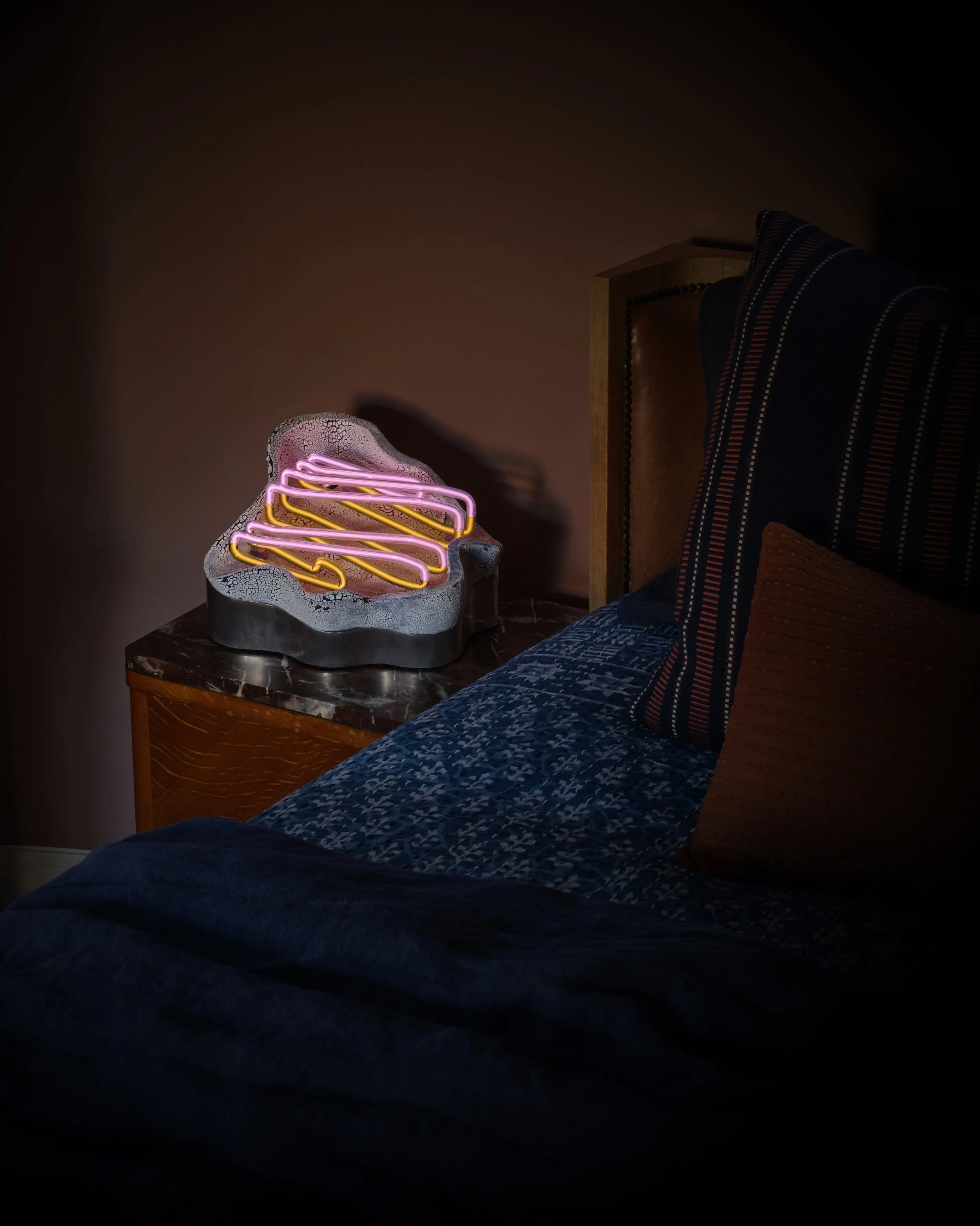 A neon sign shaped like a bear head with pink and yellow lights, placed on a bedside table next to a bed with pillows and a blue patterned blanket.