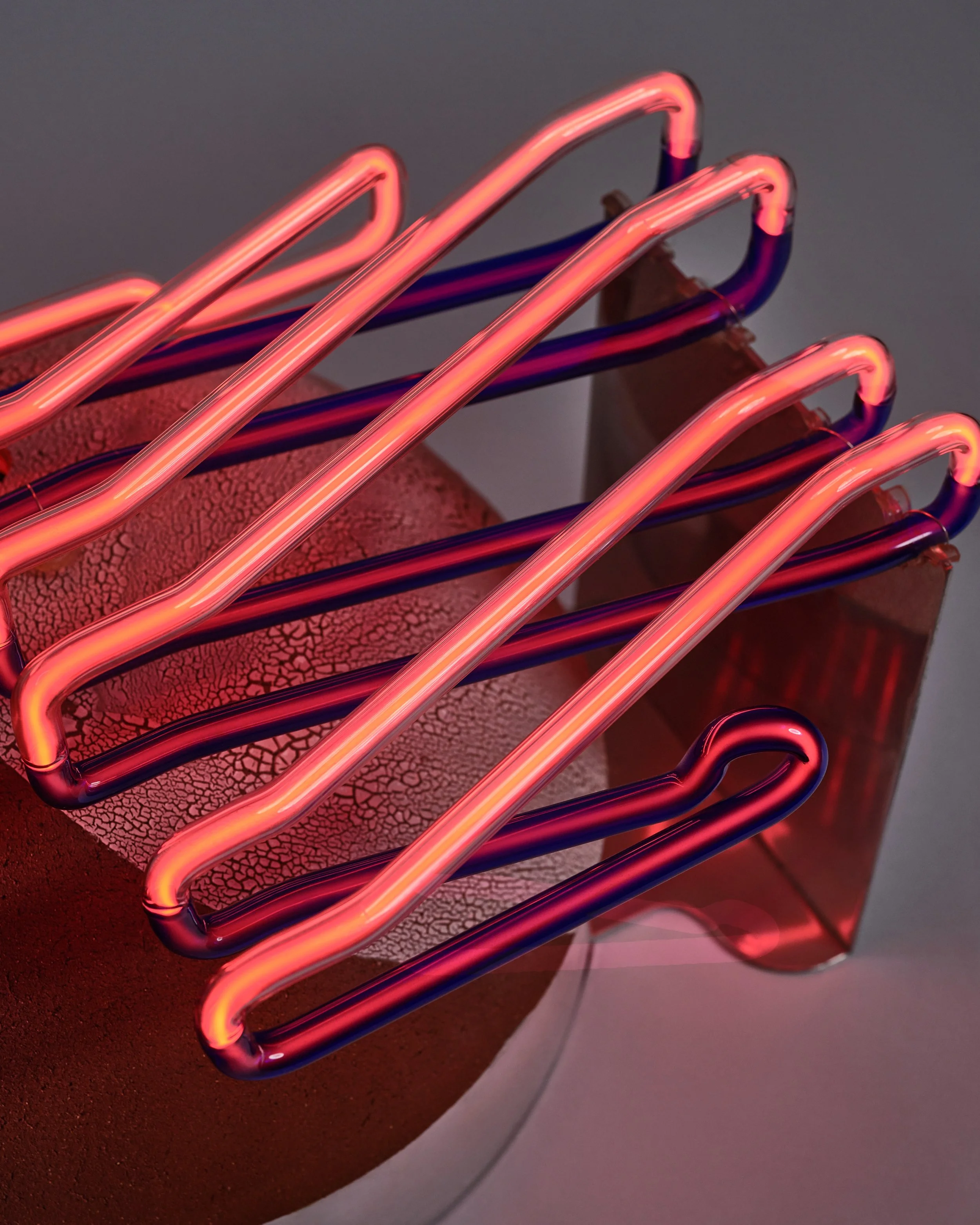 Close-up of a neon light sculpture with pink and purple glowing tubes arranged in parallel on a textured, cracked surface.