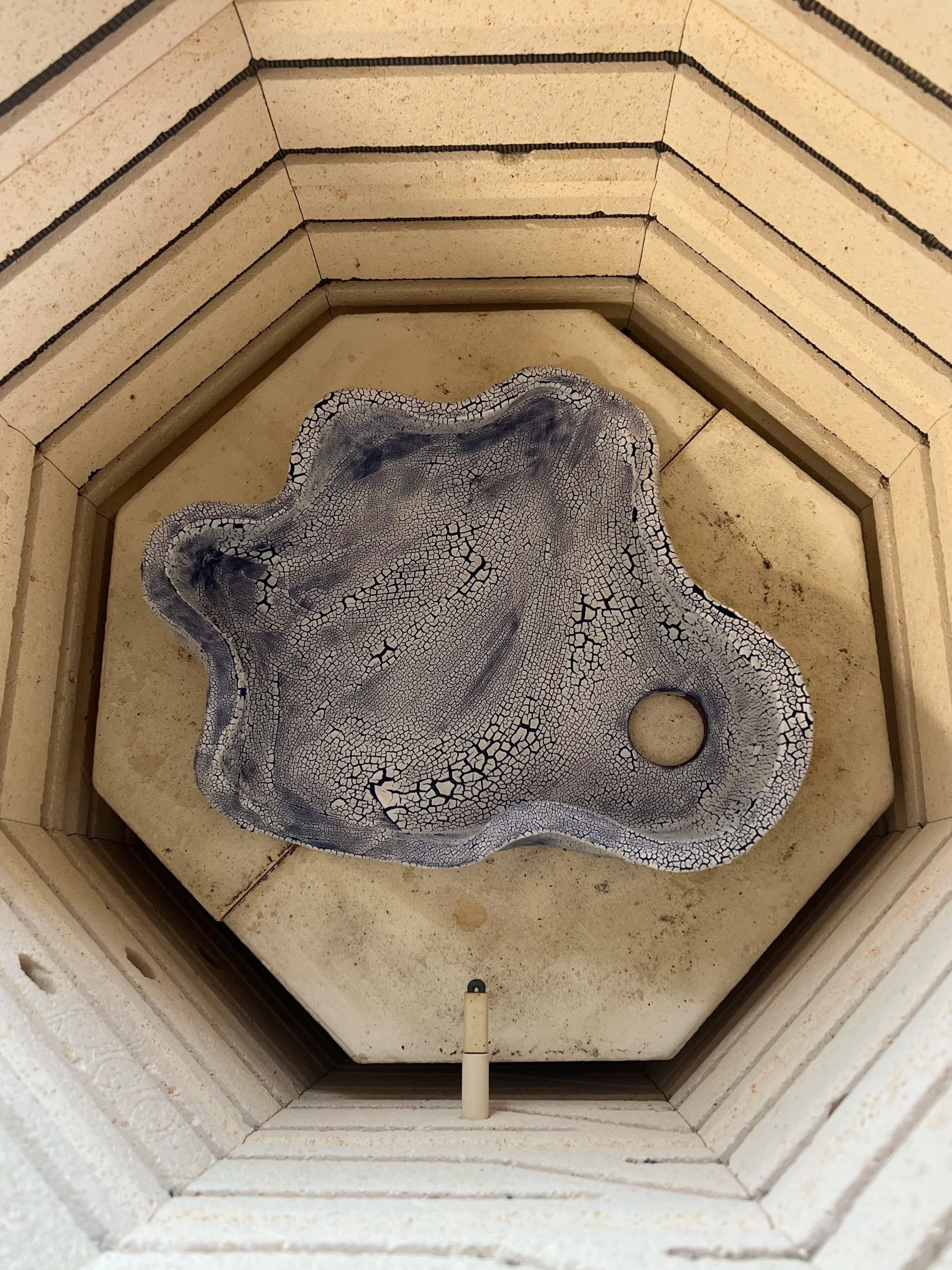 A cracked, weathered ceramic or stone decorative piece with a hole in one corner, placed inside a multi-layered, octagonal wooden or concrete structure with concentric rings.