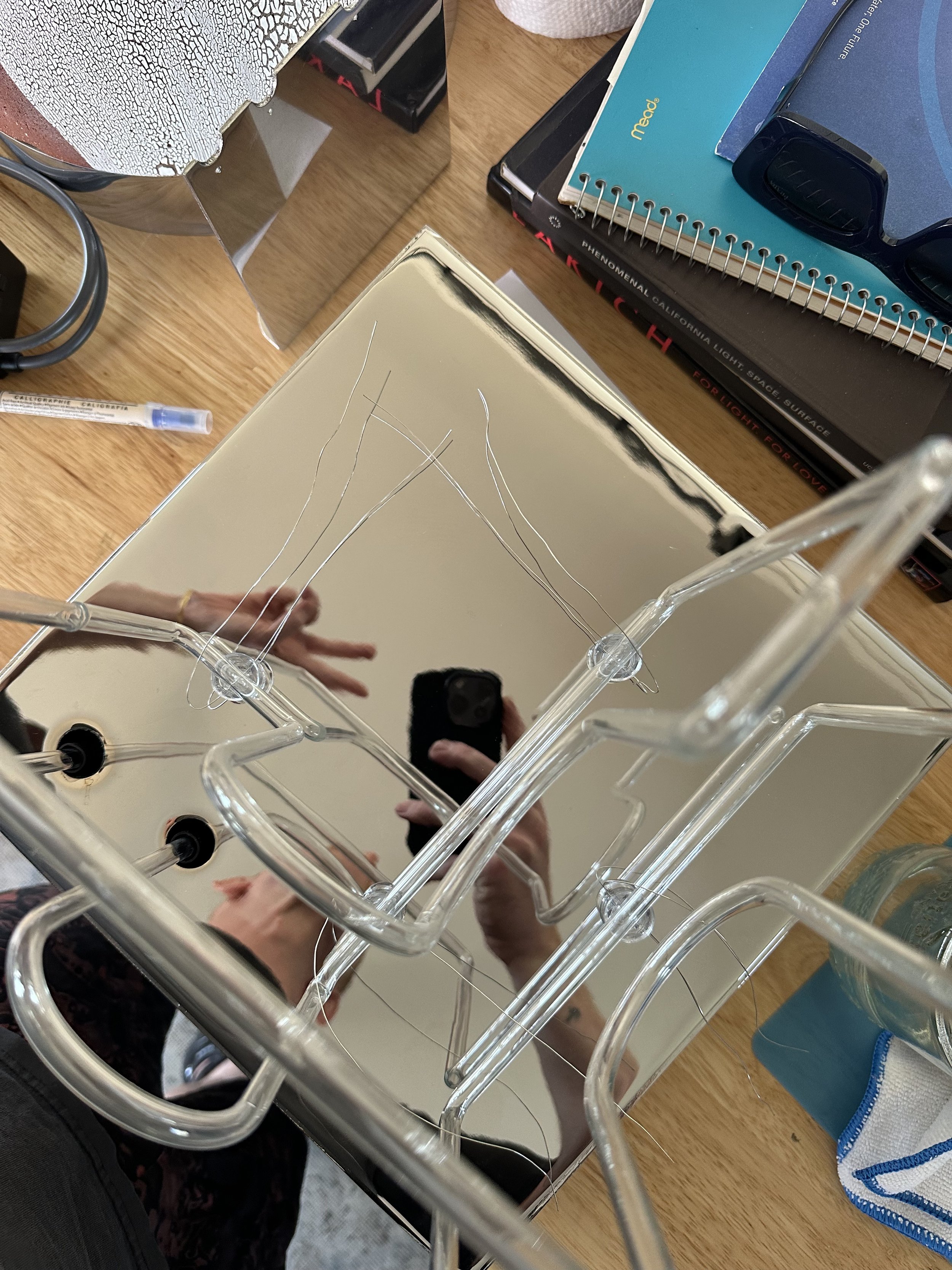 A mirror reflecting a person's hand, a smartphone, and various objects on a cluttered desk, including notebooks, a water bottle, and pens.