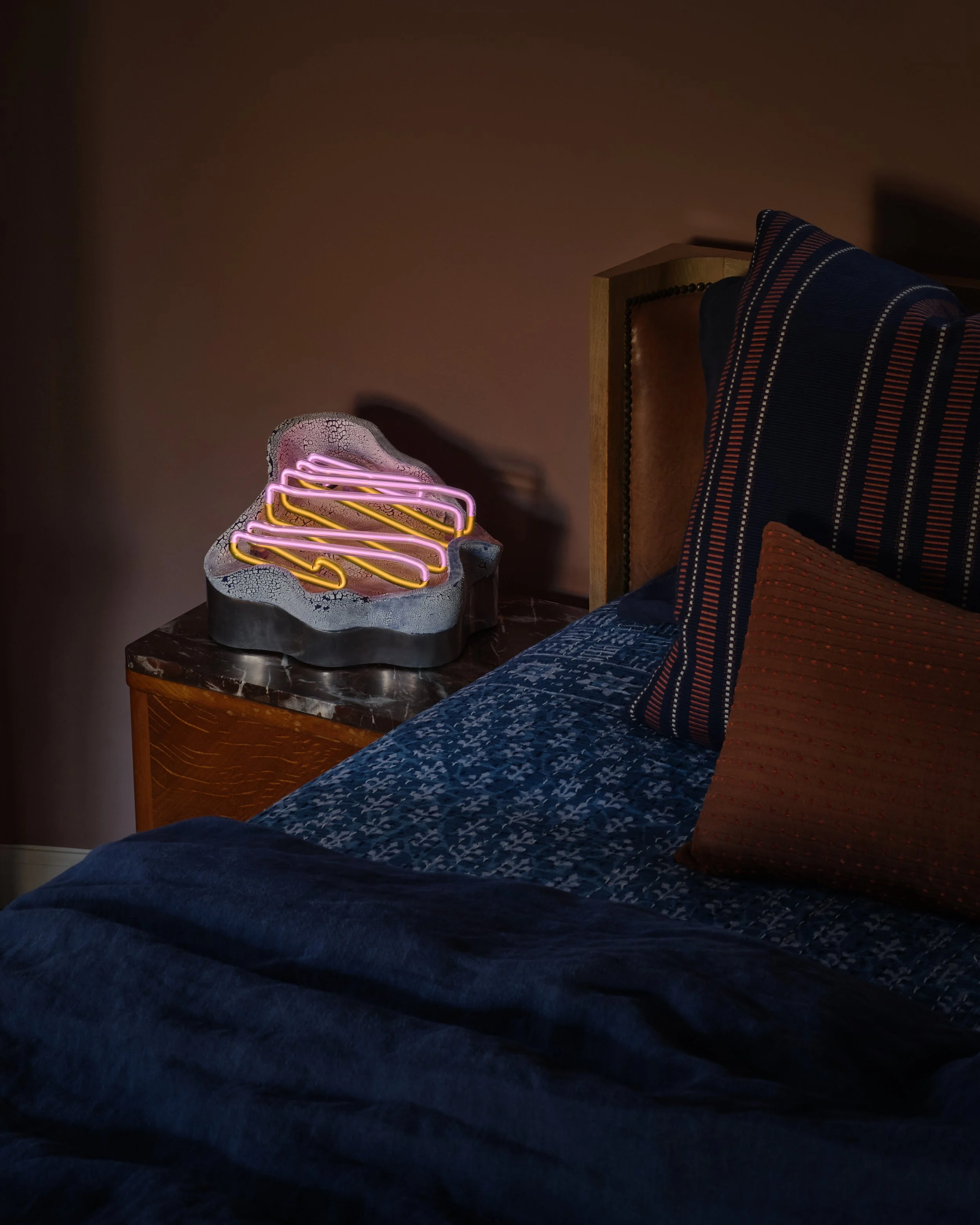 A neon pink and yellow sign shaped like a geode on a marble-top bedside table next to a bed with dark blue and striped pillows.