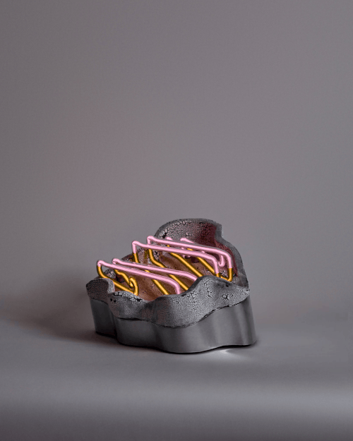 A donut with pink and yellow neon icing decorations, placed in a black irregular-shaped container against a plain gray background.