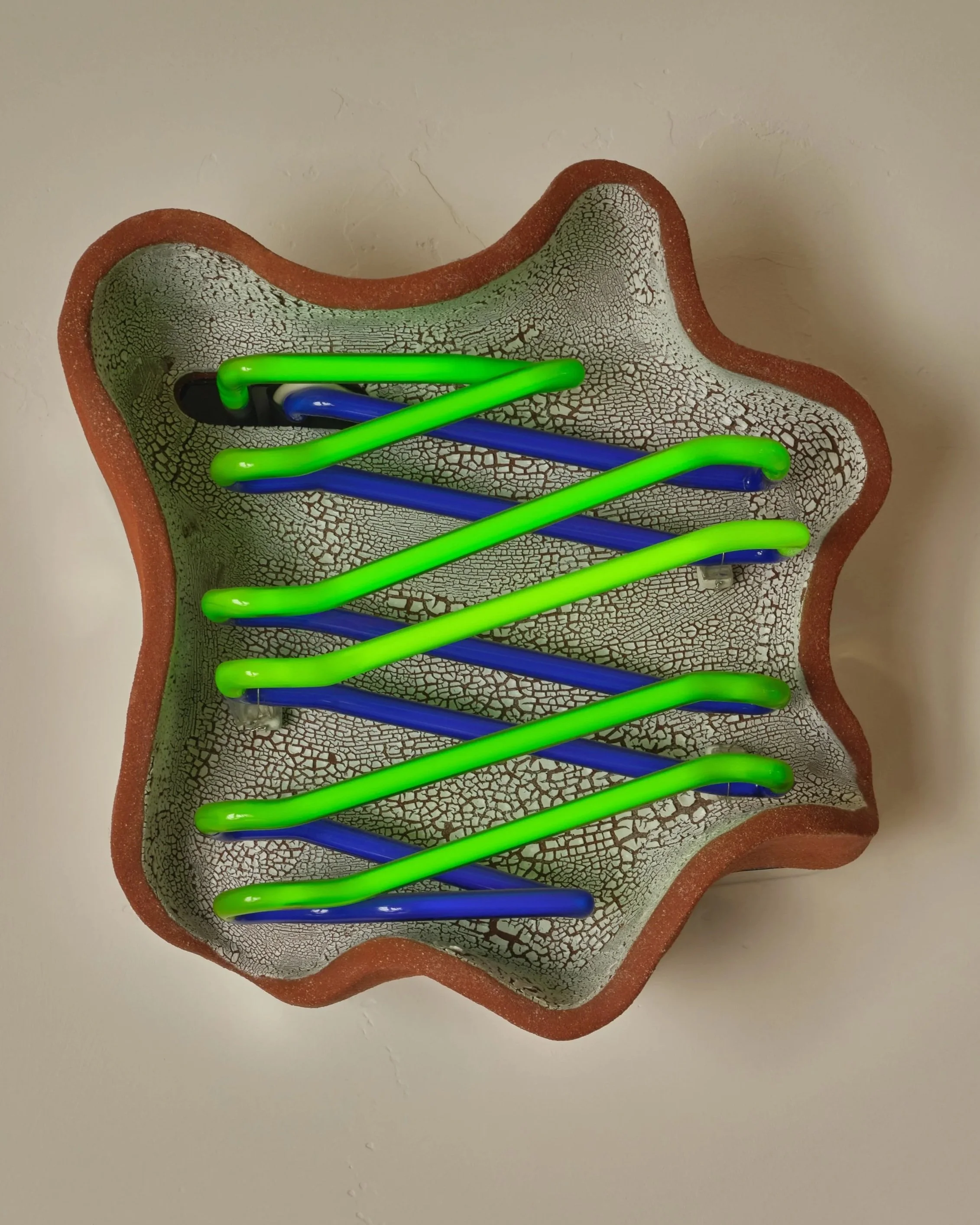 Decorative ceramic bowl with a cracked glaze pattern and a wavy edge, holding a green and blue zigzag neon light installation.