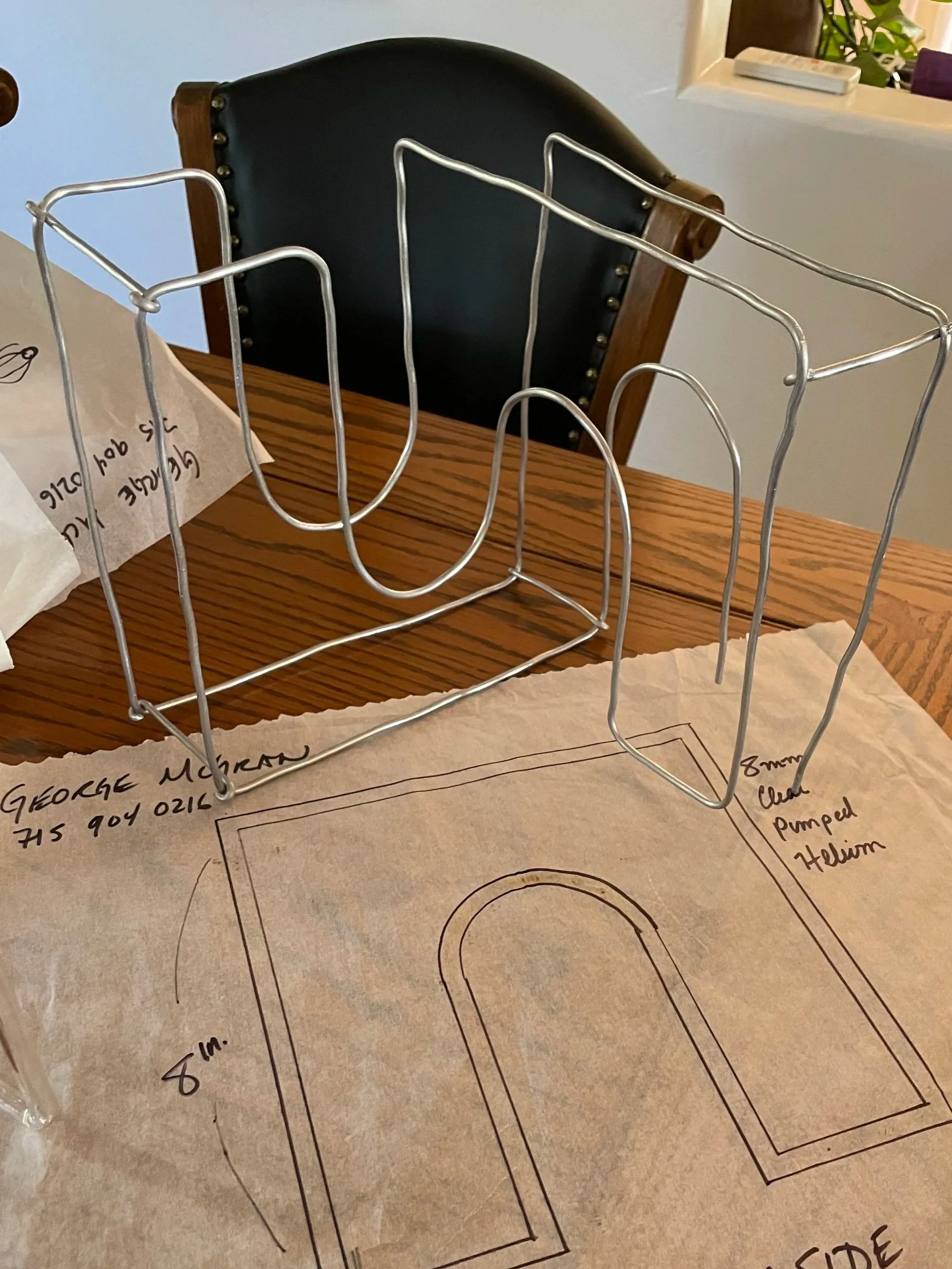 Metal wire model of an infinity arch glyph standing upright on a paper with drafting lines, on a wooden table with a black chair in the background.