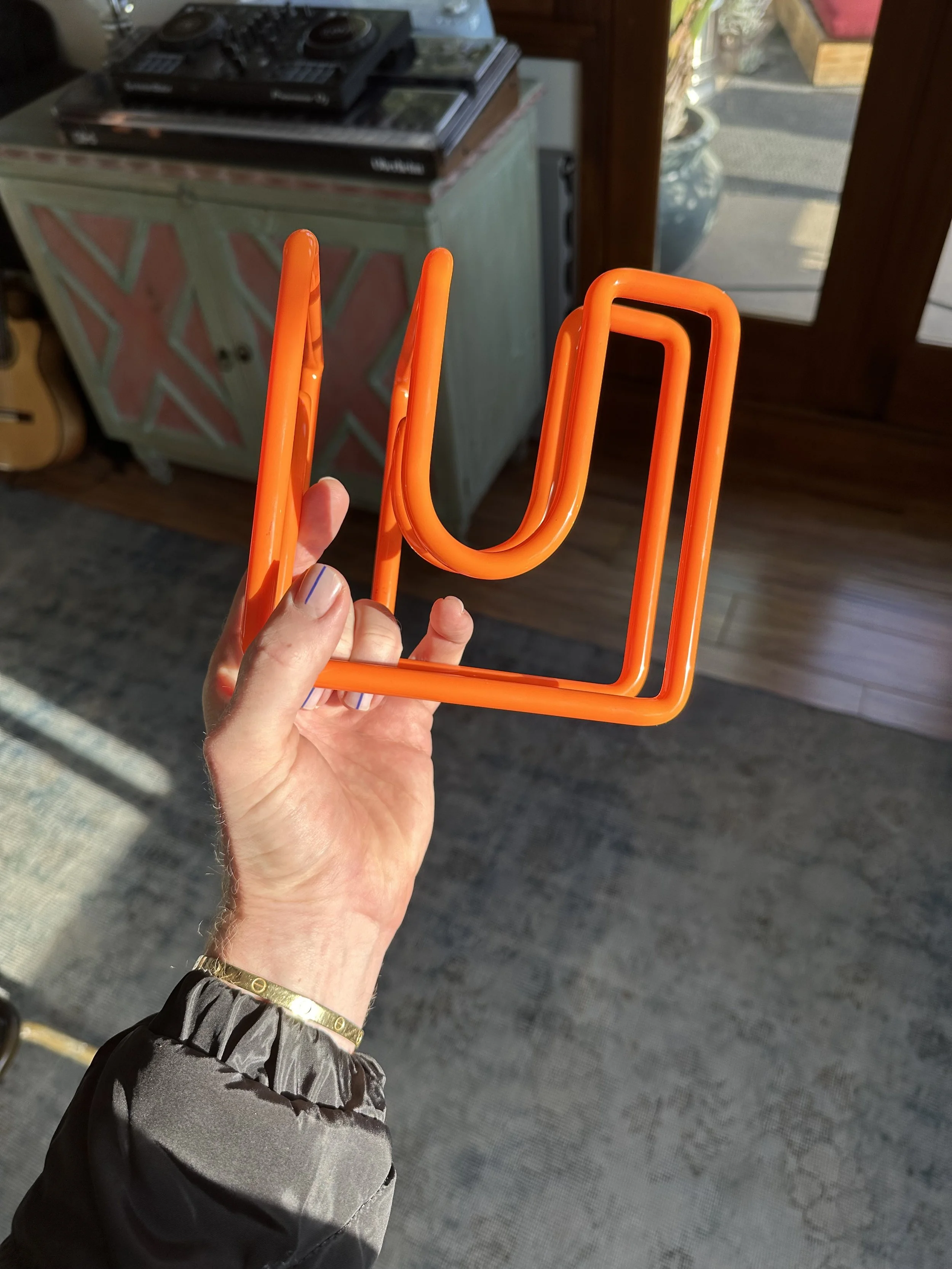 Hand holding a bright orange neon sign shaped like a letter U with a square outline. In the background, there is a green piece of furniture with a geometric pattern and a DJ set on top of it. The setting appears to be inside a room with a wooden door or window leading outside.
