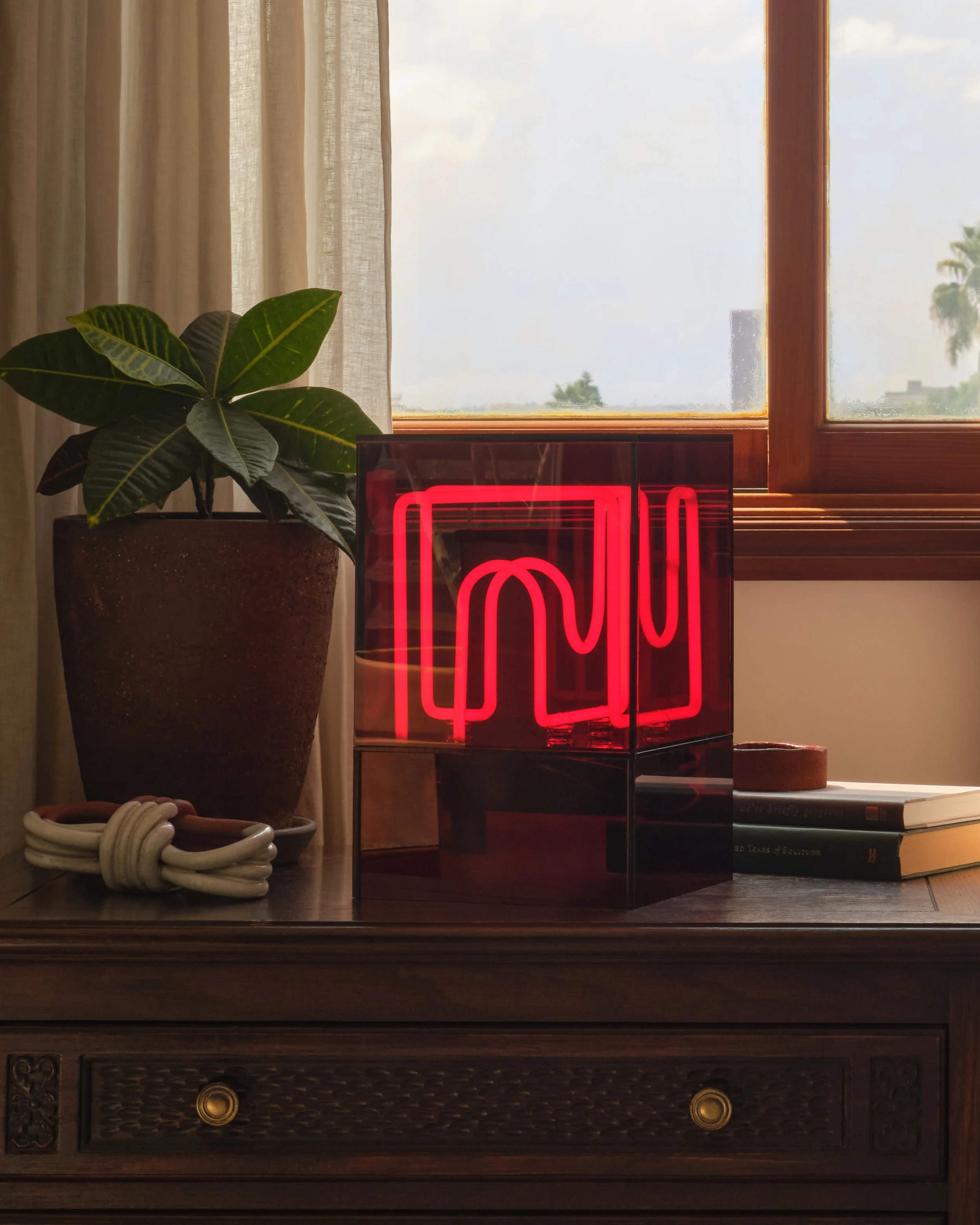 A windowsill with a potted plant, a neon red art piece, and some books with a curtain and window in the background.